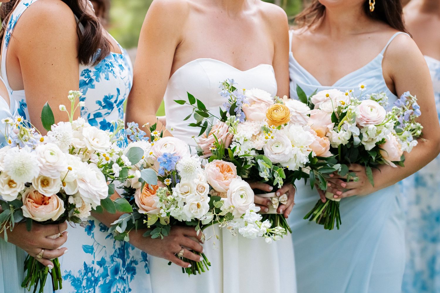 Bride and bridesmaids in blue dresses hold stunning white, peach, and blush floral wedding bouquets.