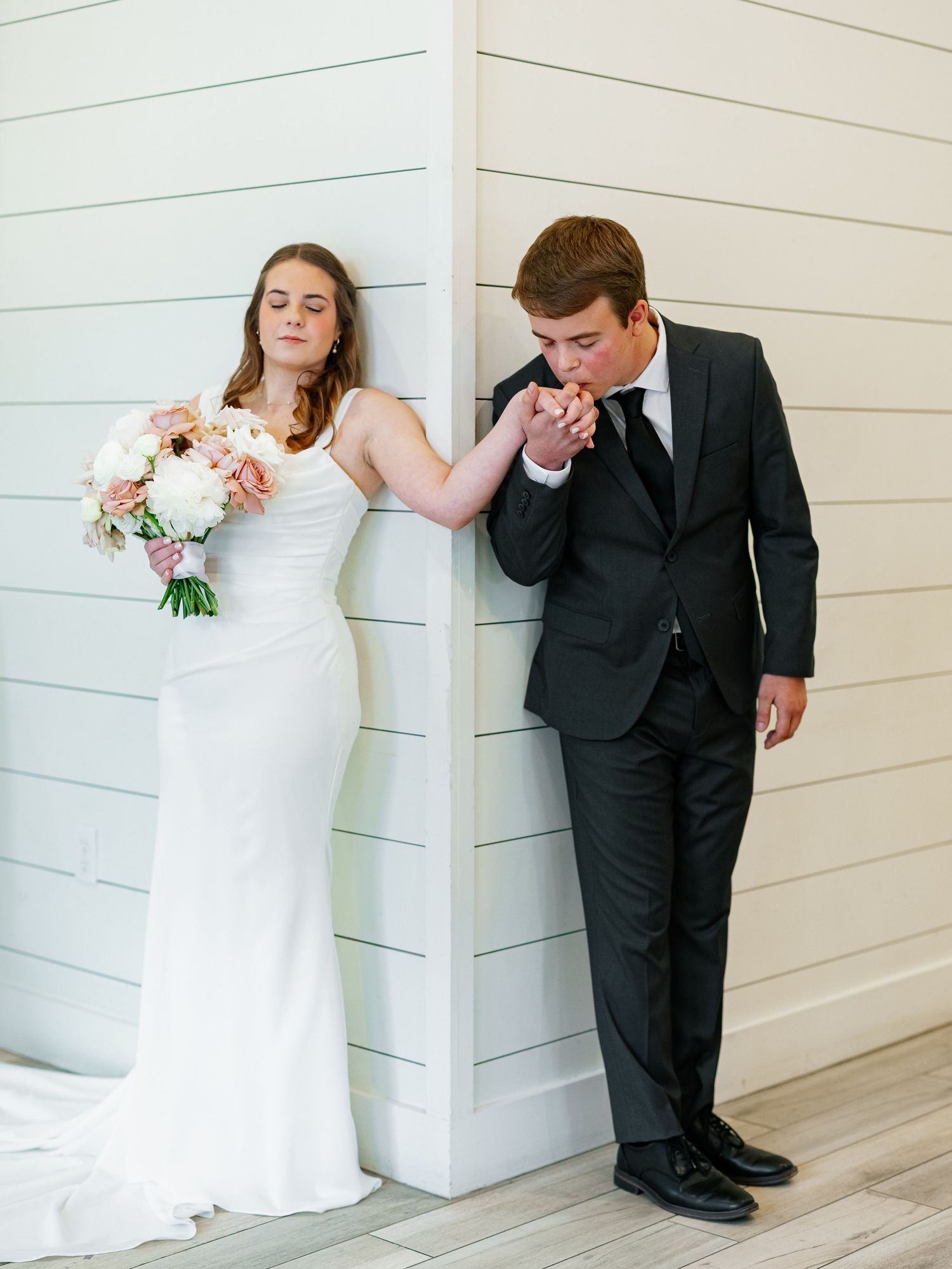 Groom in black suit kissing bride's hand as she leans against white shiplap wall holding bouquet.