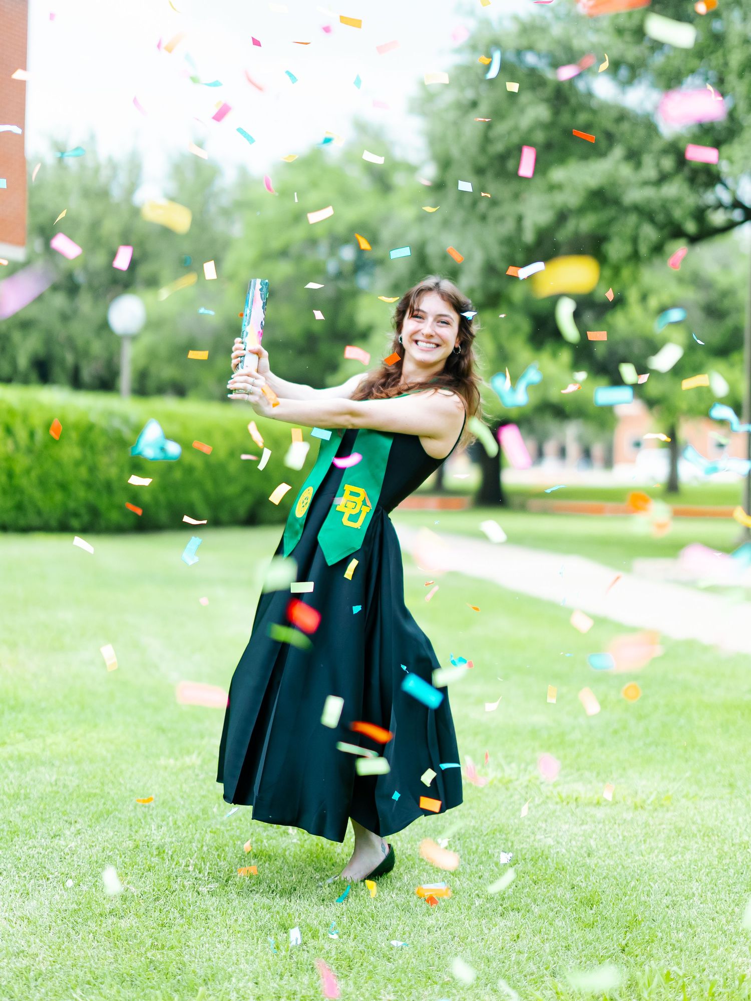 Joyful Baylor University graduate celebrating with confetti cannon on green campus lawn.