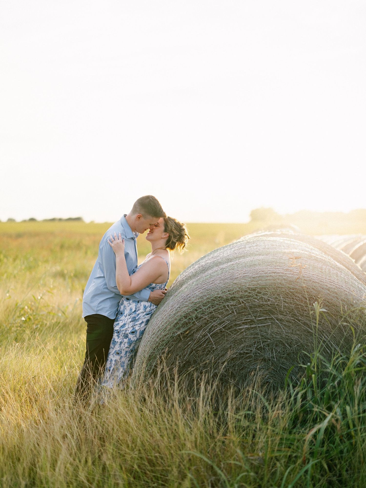 Couple sharing a romantic kiss beside a round hay bale in a golden sunlit field during sunset.