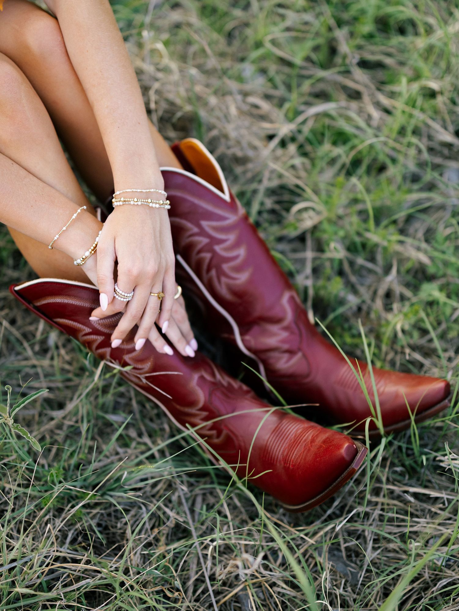 Woman wearing red cowboy boots in grass, adorned with gold bracelets and rings, showcasing western fashion style.