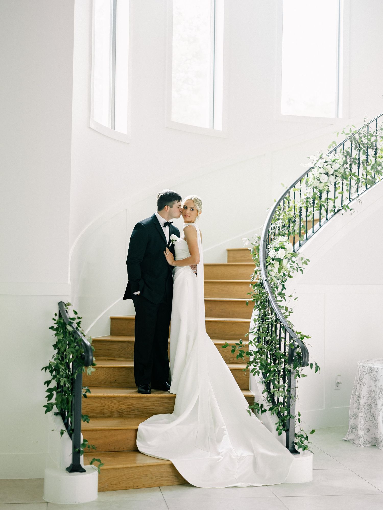 Bride and groom share a kiss on elegant white staircase adorned with greenery in a bright wedding venue.