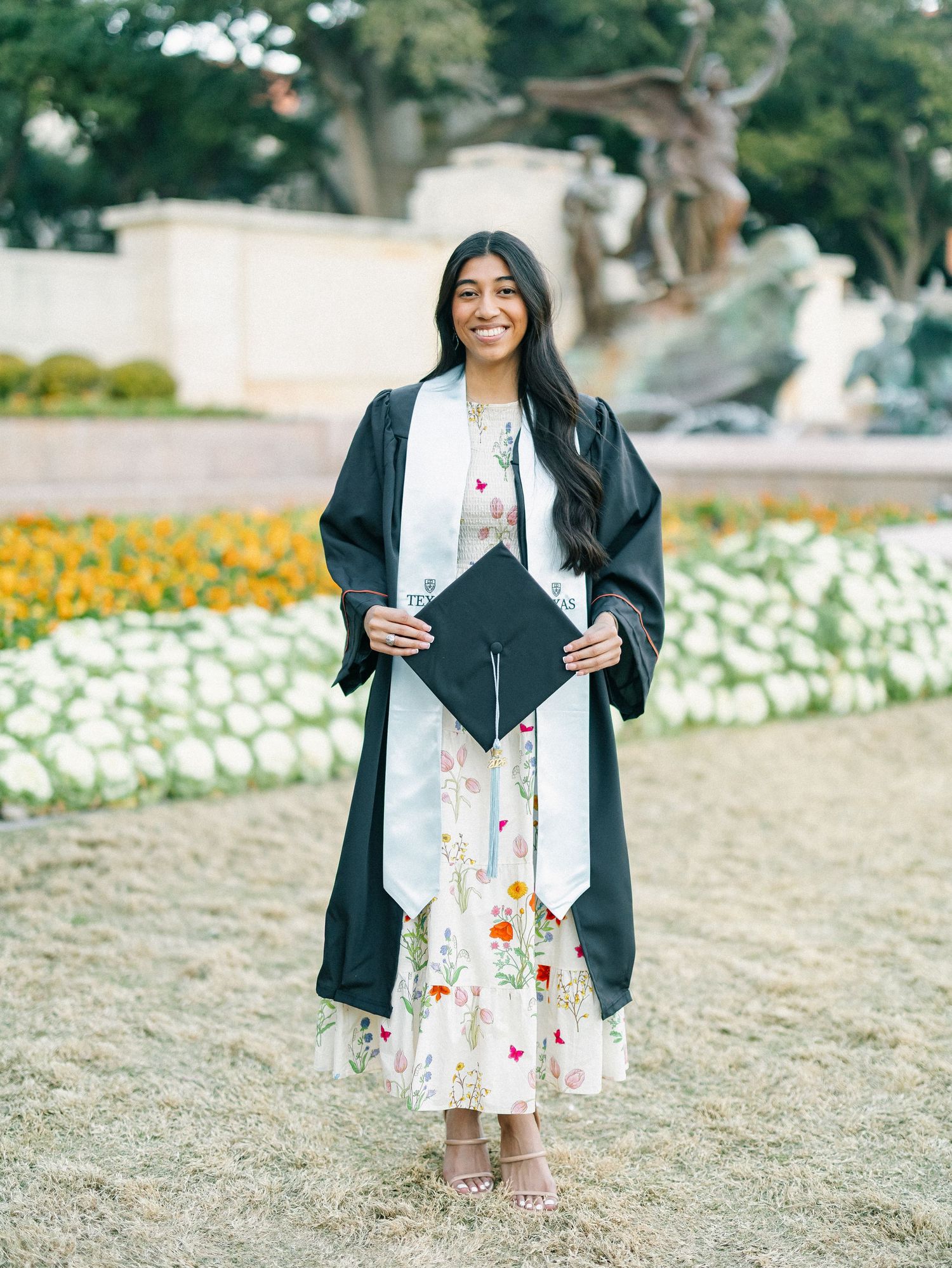 Smiling graduate in black cap and gown holding diploma, standing in front of colorful flower garden.