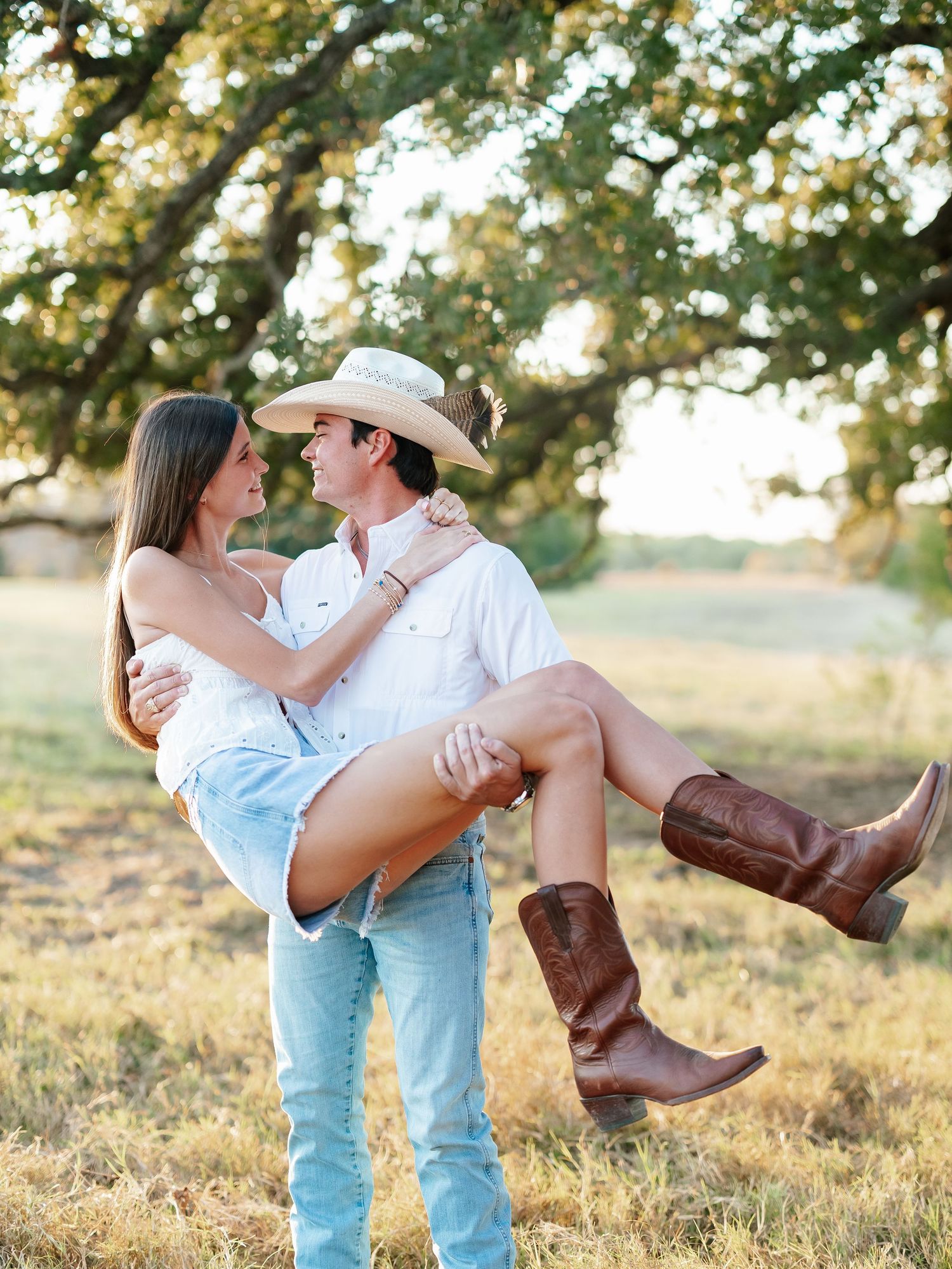 A cowboy in a white shirt and hat lifts a smiling woman in a sunny green field.