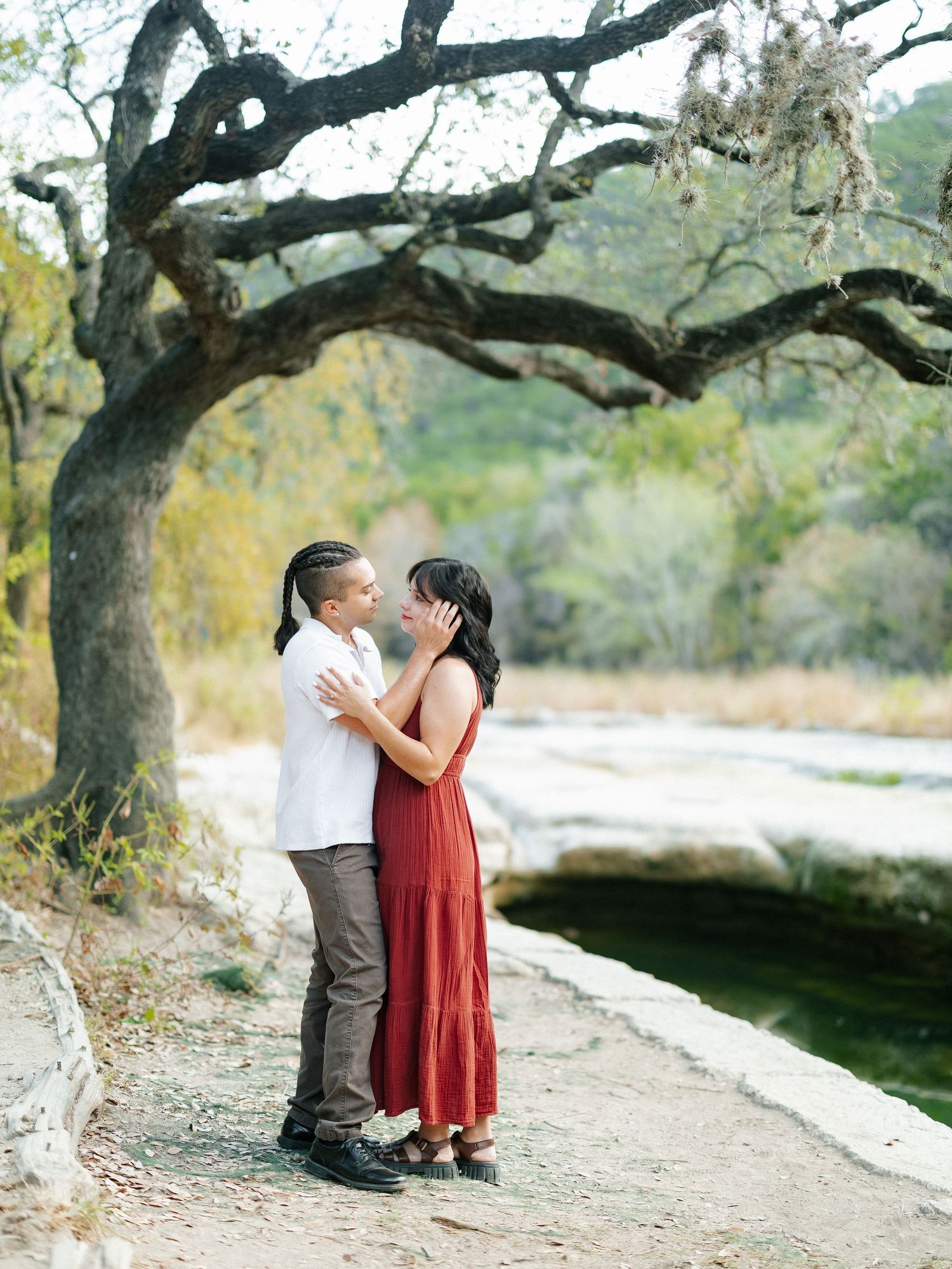 Couple sharing a romantic moment under a sprawling oak tree by a rocky creek in nature.
