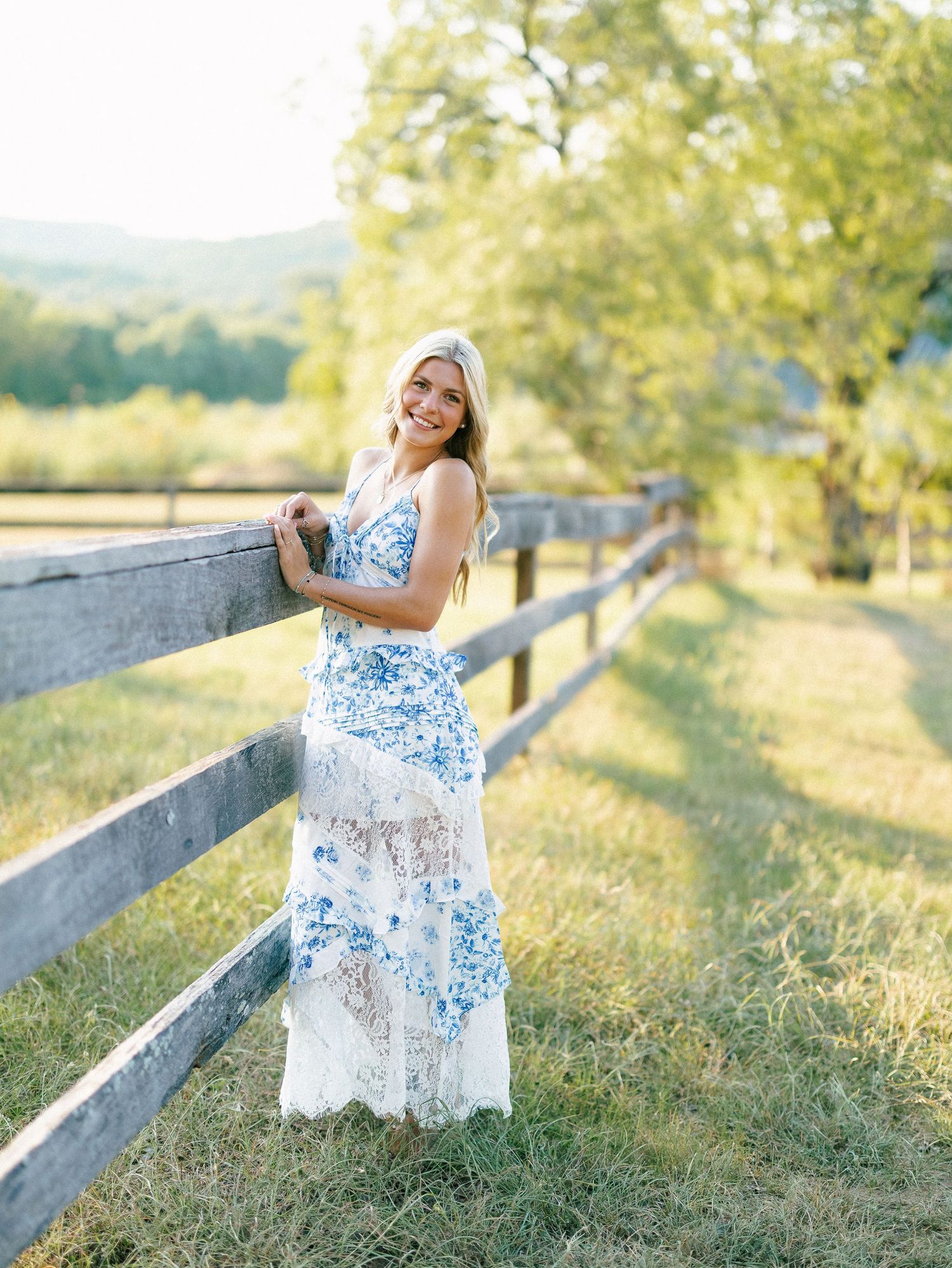 Young woman in blue floral maxi dress smiling by rustic wooden fence in sunny countryside setting.