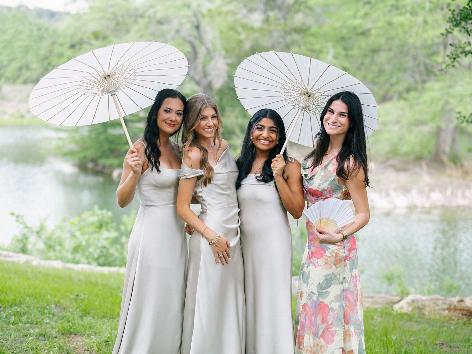 Four bridesmaids in silver gowns holding white parasols smile outdoors near a river at a wedding.
