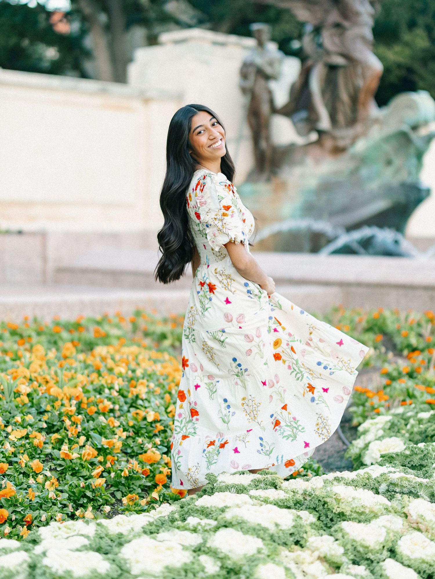 Woman in floral maxi dress twirling in a garden with orange and white flowers and a fountain backdrop.