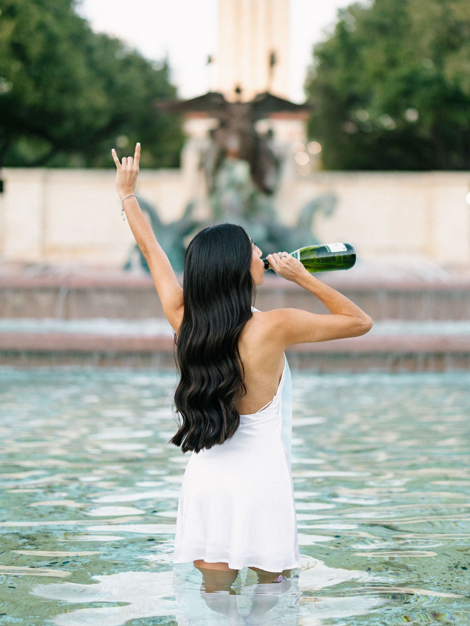 Woman in white dress standing in fountain, raising hand and drinking from champagne bottle.