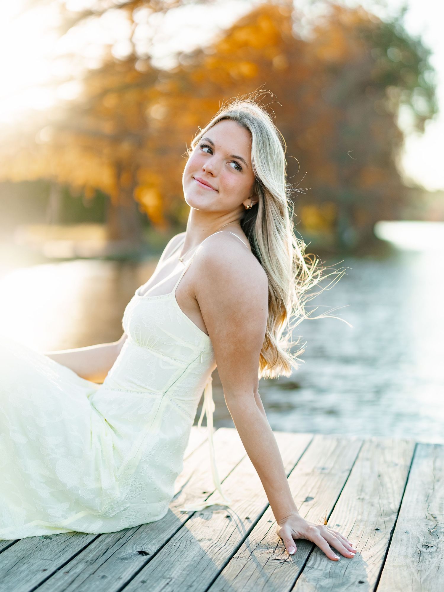 Blonde woman in white dress sitting on wooden dock by lake with autumn foliage in golden sunlight.
