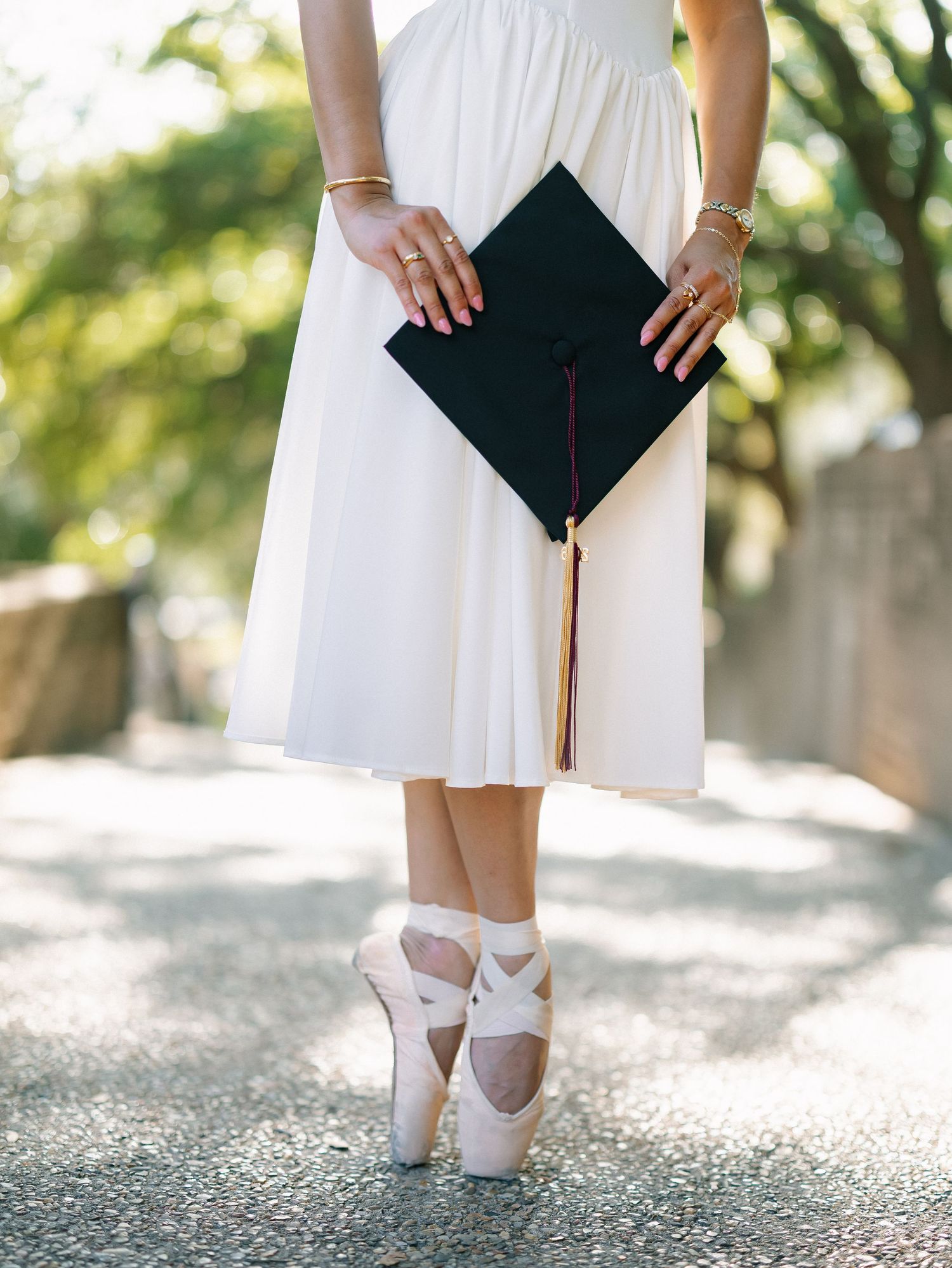 Woman in white dress and ballet heels holding a black graduation cap with gold tassel outdoors.
