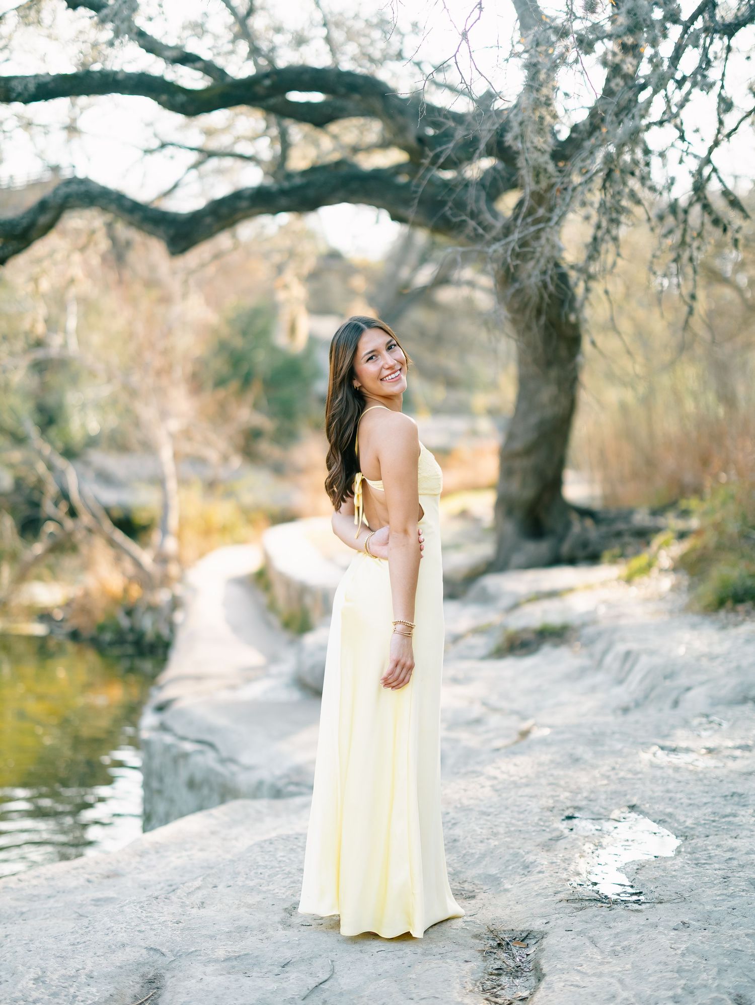Young woman in yellow maxi dress smiling outdoors near rocky creek with oak trees in natural light.