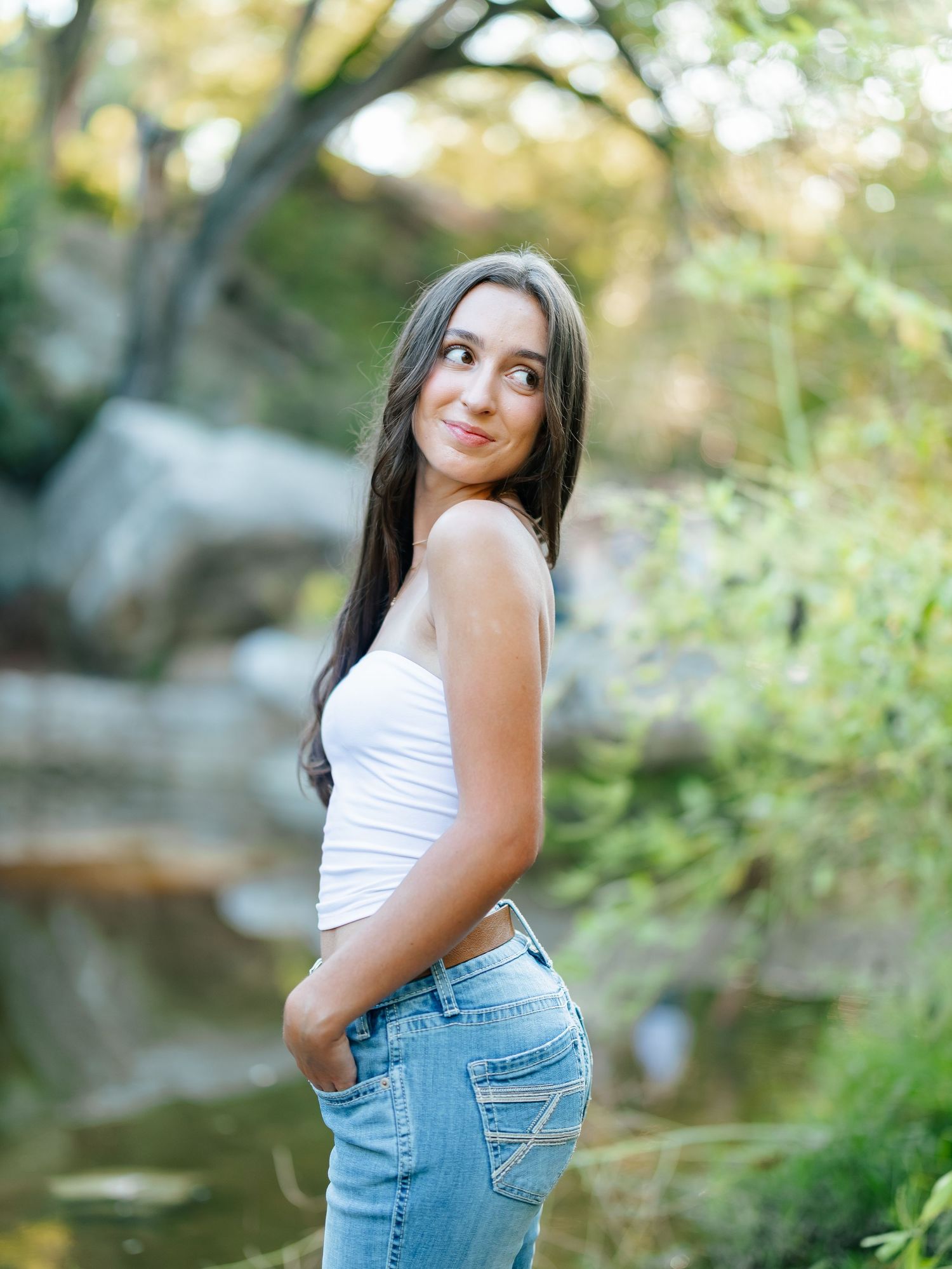 Young woman in white tank top and light jeans poses outdoors near a rocky creek surrounded by lush greenery.