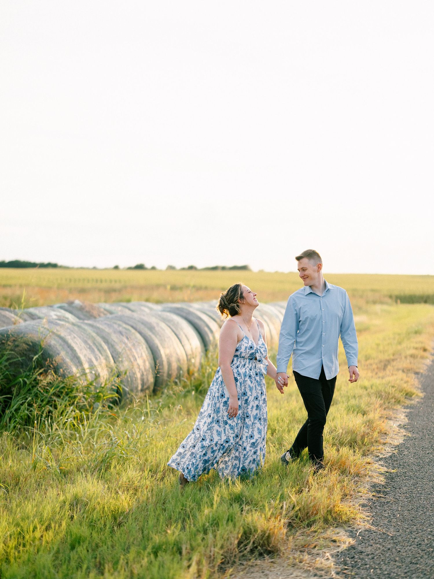 Couple holding hands walking along a rural path beside hay bales in a golden field at sunset.