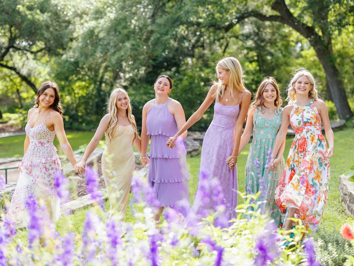 Six women in colorful floral and pastel maxi dresses hold hands in a lush garden with purple wildflowers blooming in the foreground.