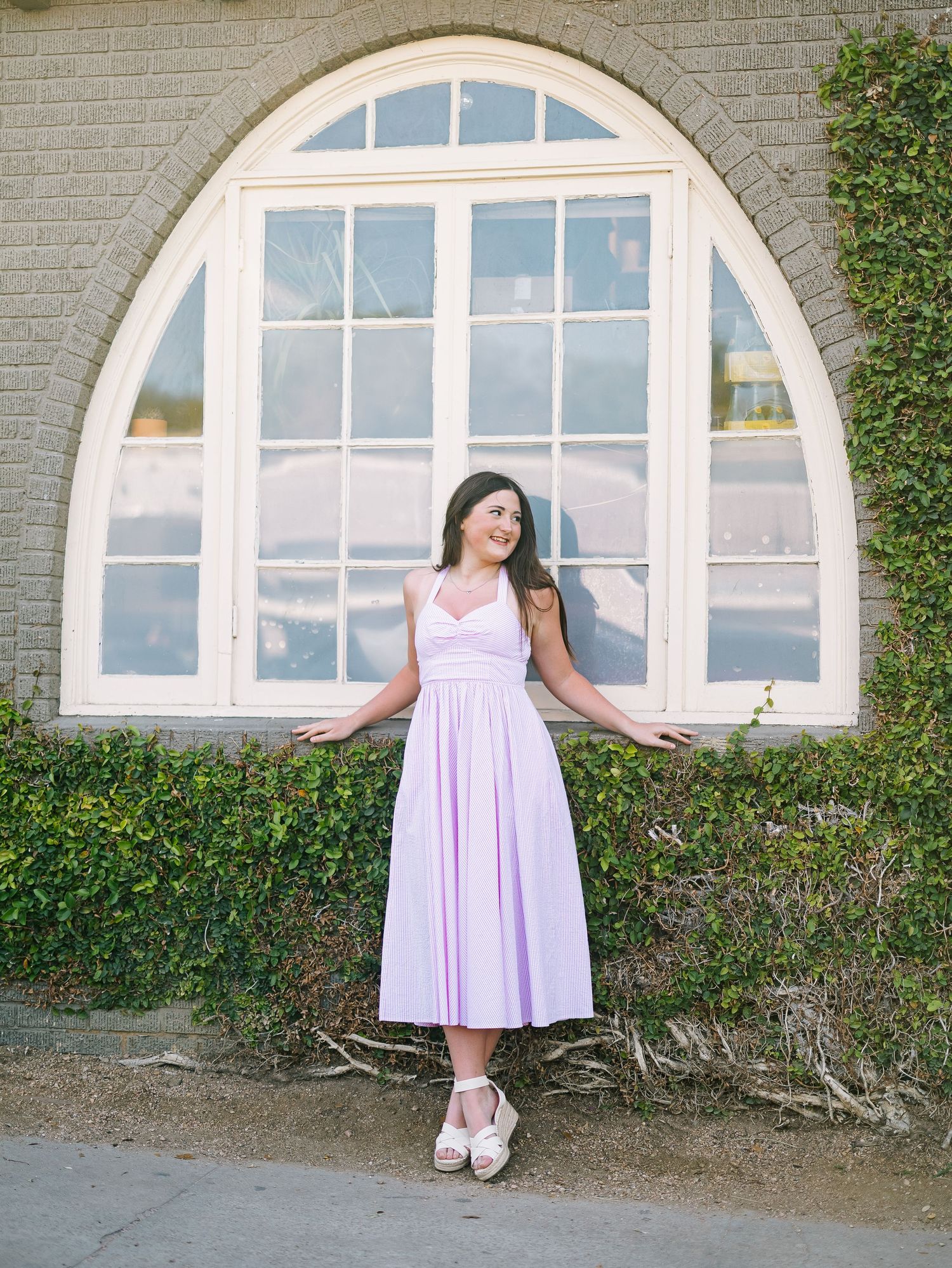 Woman in lavender midi dress posing in front of arched window with ivy-covered stone wall.