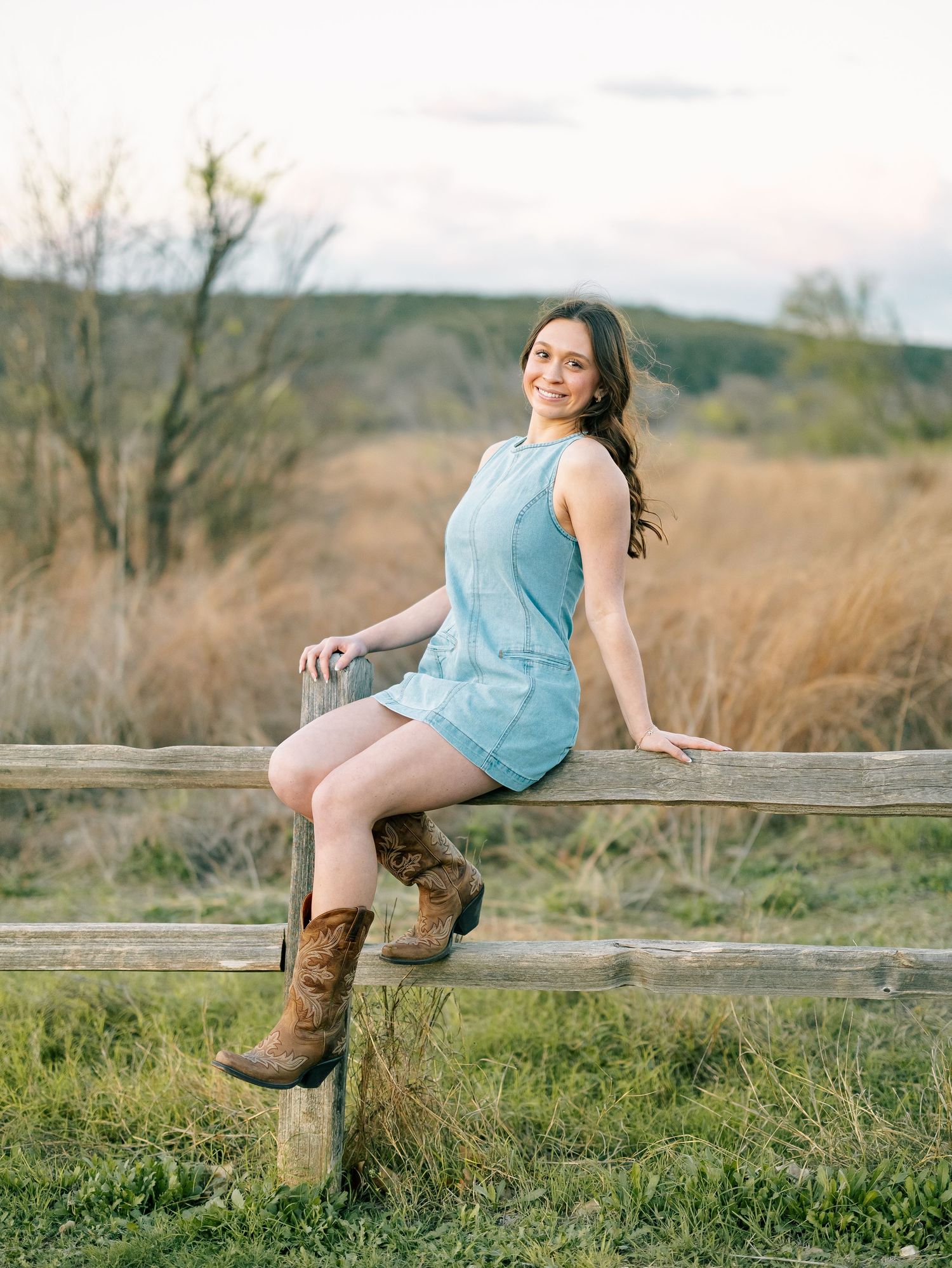 Young woman in light blue dress and cowboy boots sitting on a rustic wooden fence in a rural field.