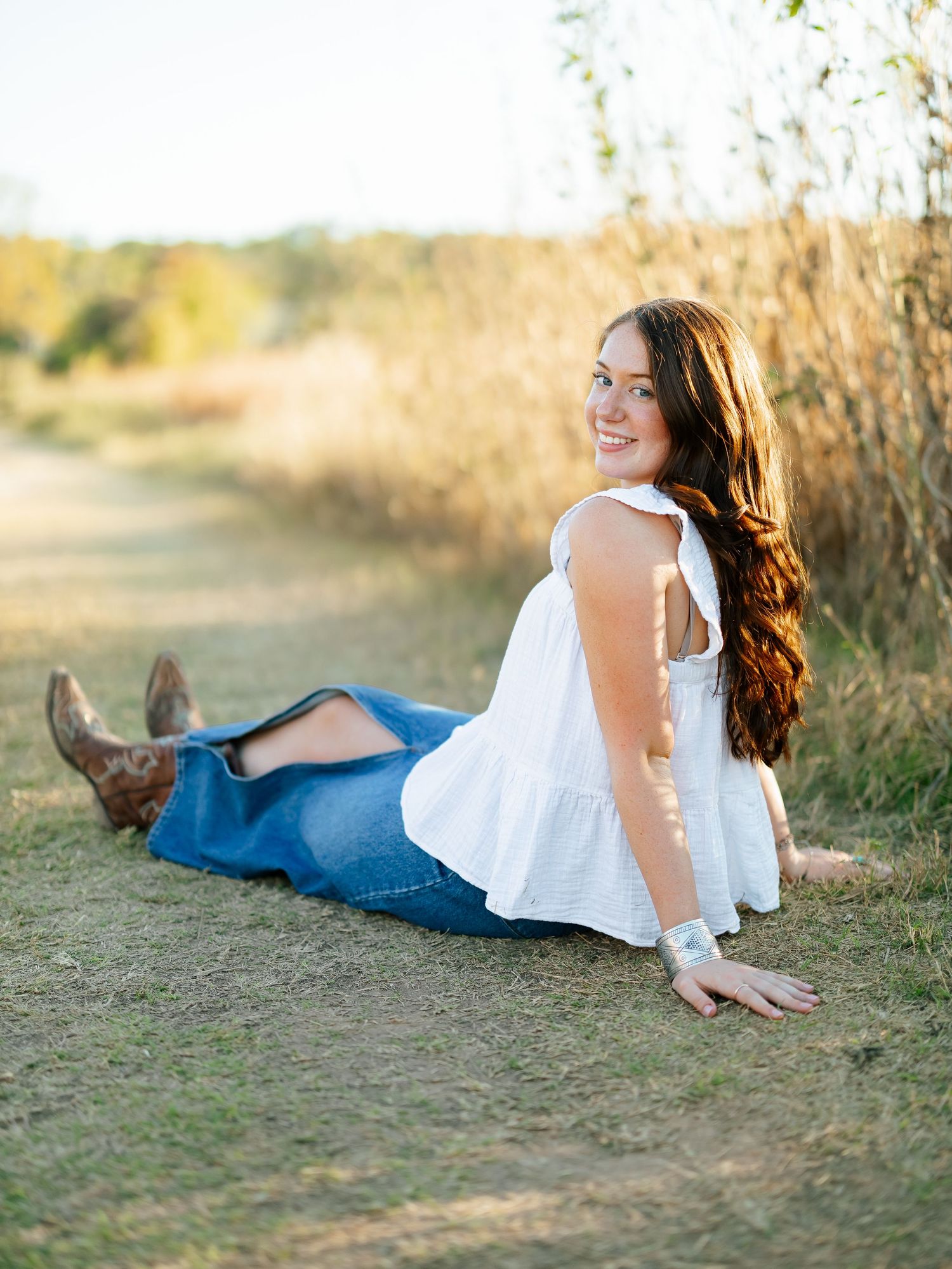 Smiling young woman in white top and denim skirt sitting on a sunlit dirt path.