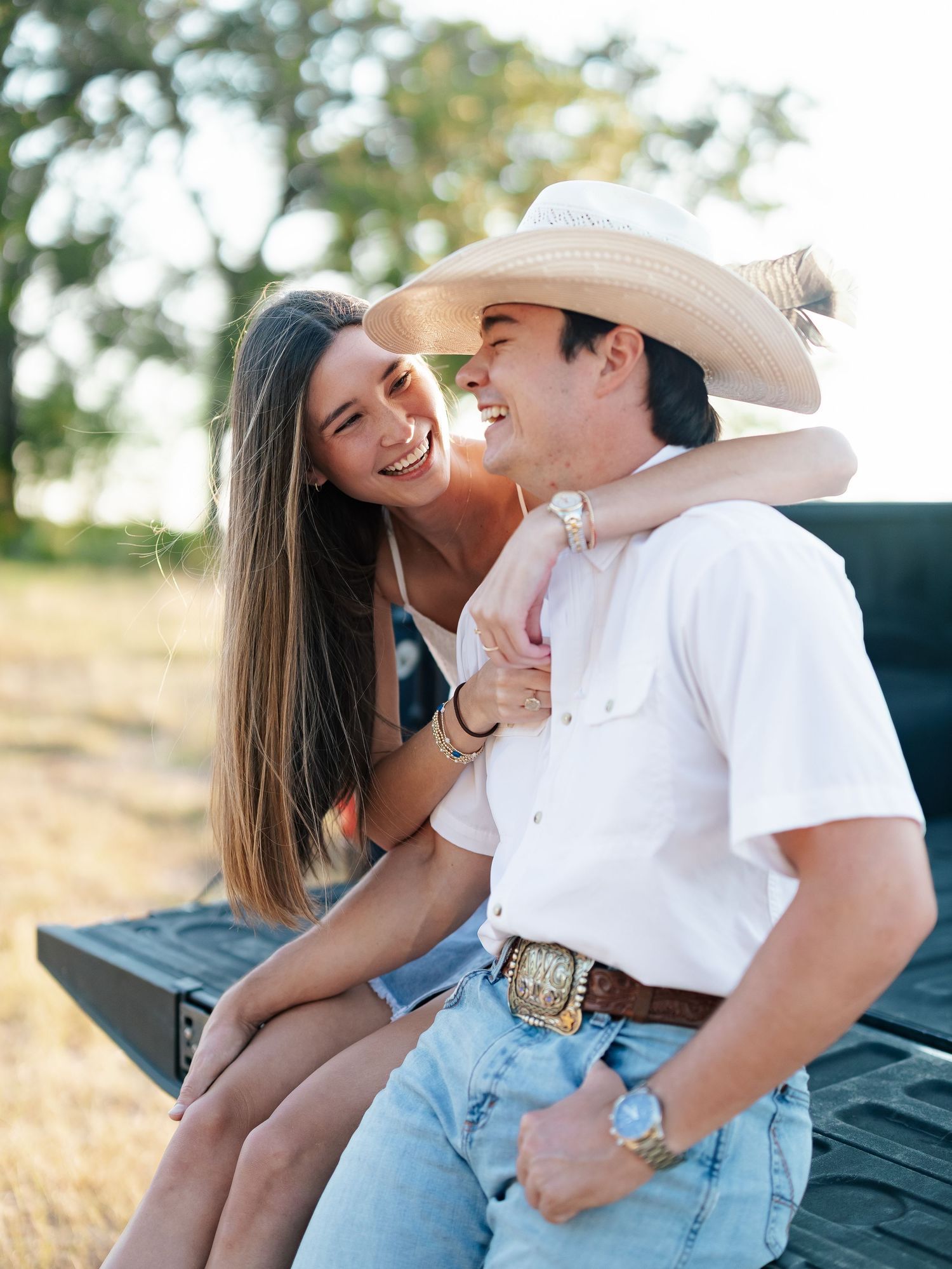 A smiling couple sits on a truck tailgate in a sunny field, man wearing cowboy hat and boots.