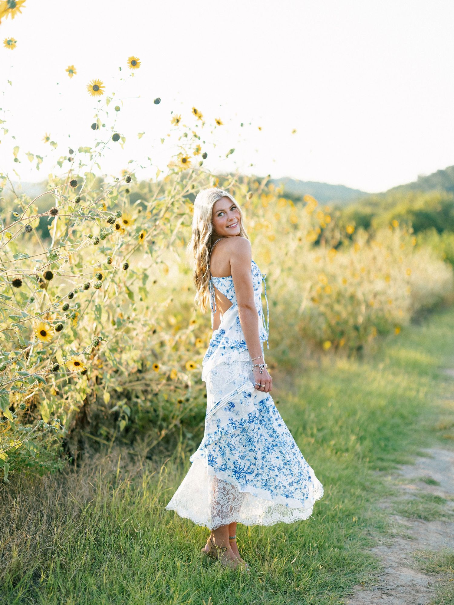 Blonde woman in blue floral lace maxi dress posing near sunflowers in a golden field at sunset.