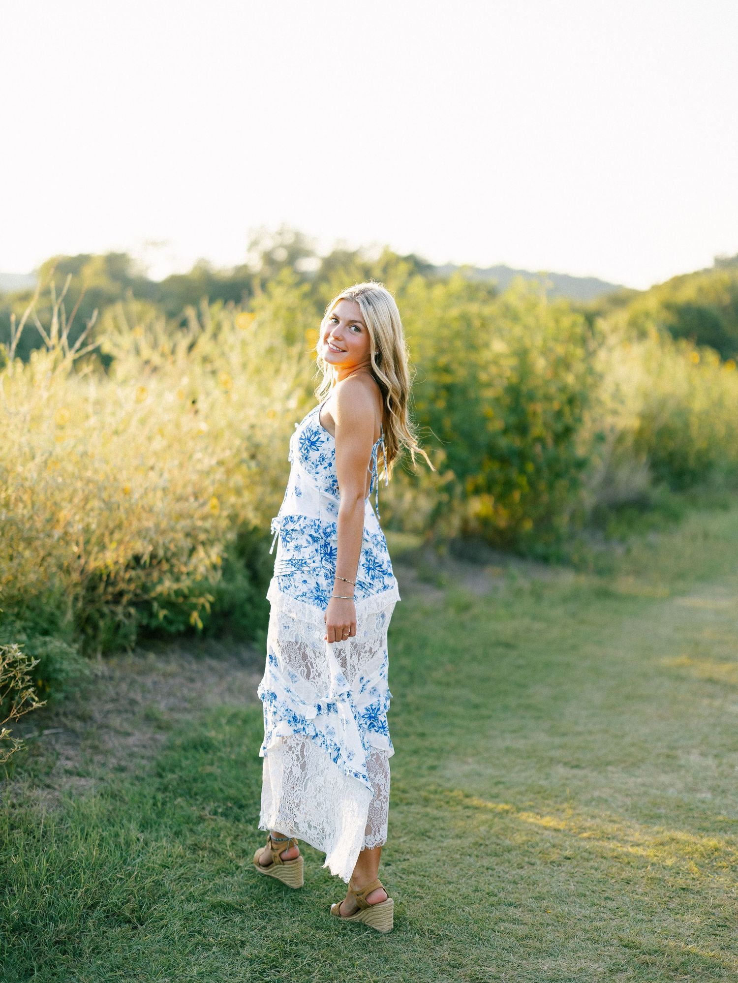 Smiling blonde woman in blue floral white maxi dress standing on green path at golden hour.