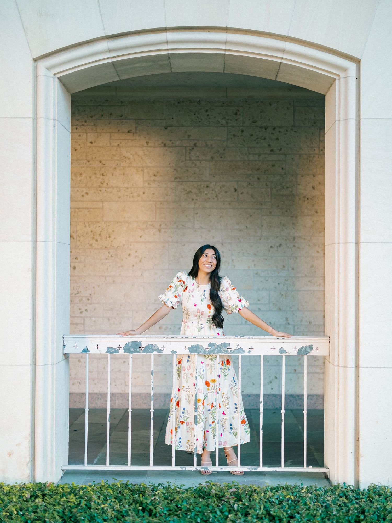 Woman in floral midi dress poses on white iron balcony framed by stone arch surrounded by lush greenery.
