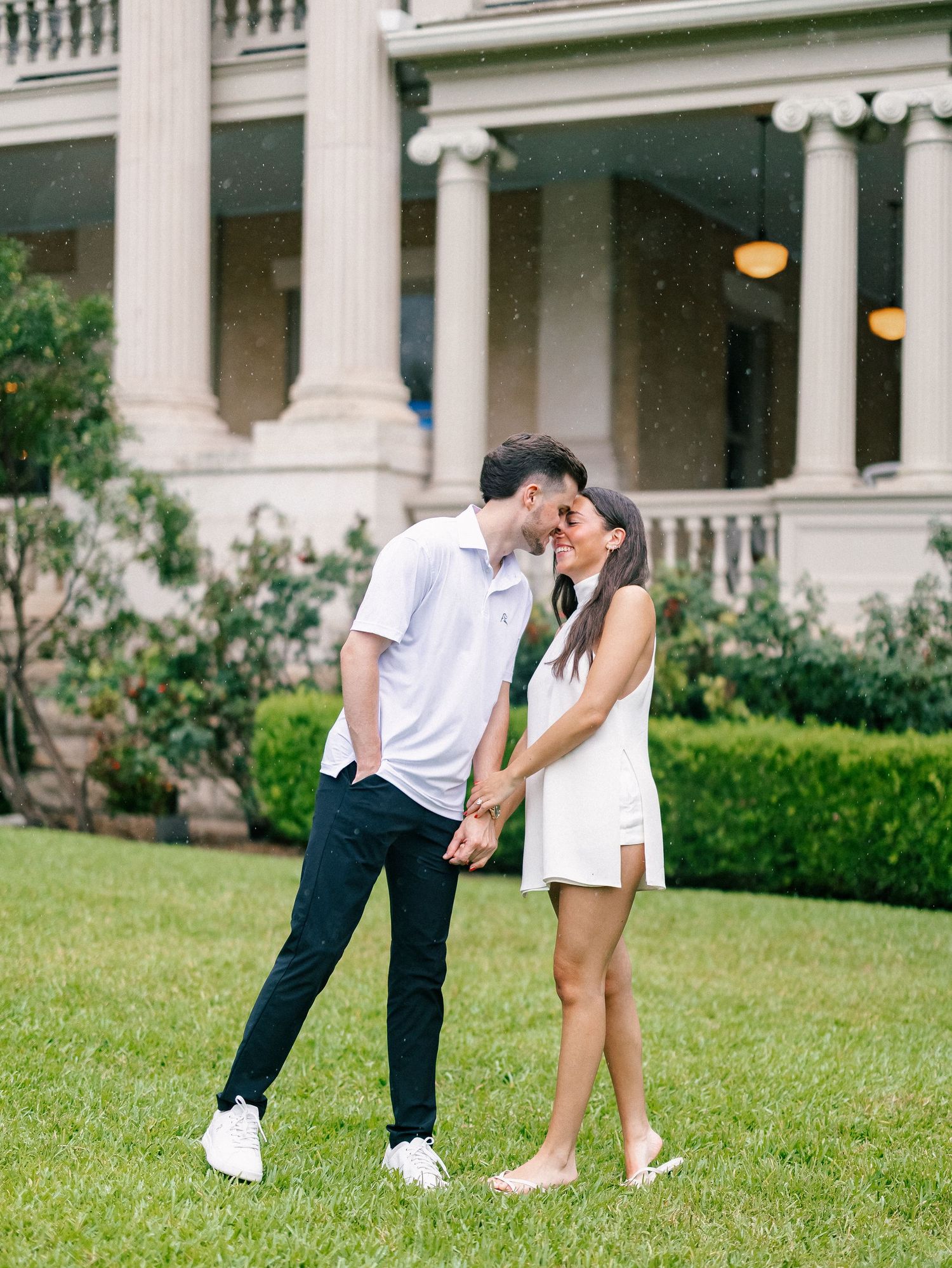 Couple holding hands and touching foreheads on lush lawn before a grand columned mansion.