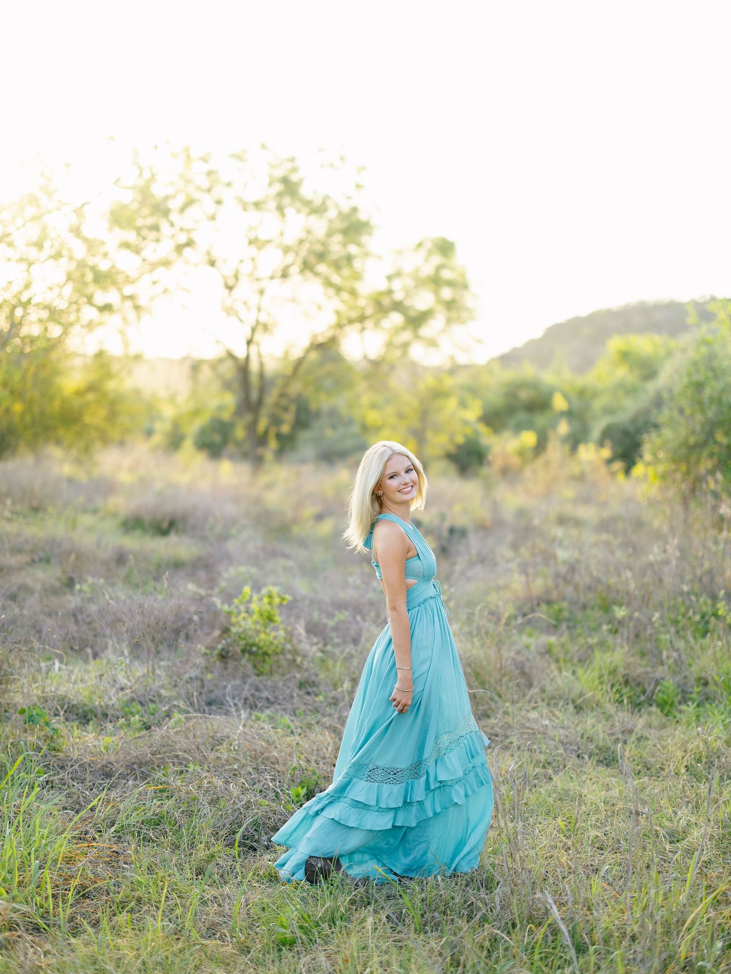 Blonde woman in flowing teal maxi dress walking through sunlit meadow.