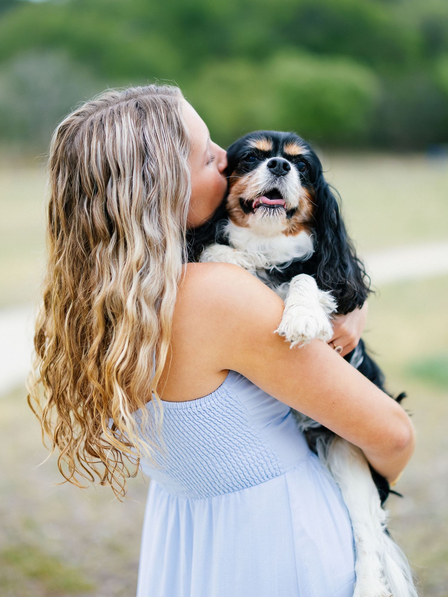 A young woman with long wavy blonde hair in a light blue dress kisses her Cavalier King Charles Spaniel outdoors.