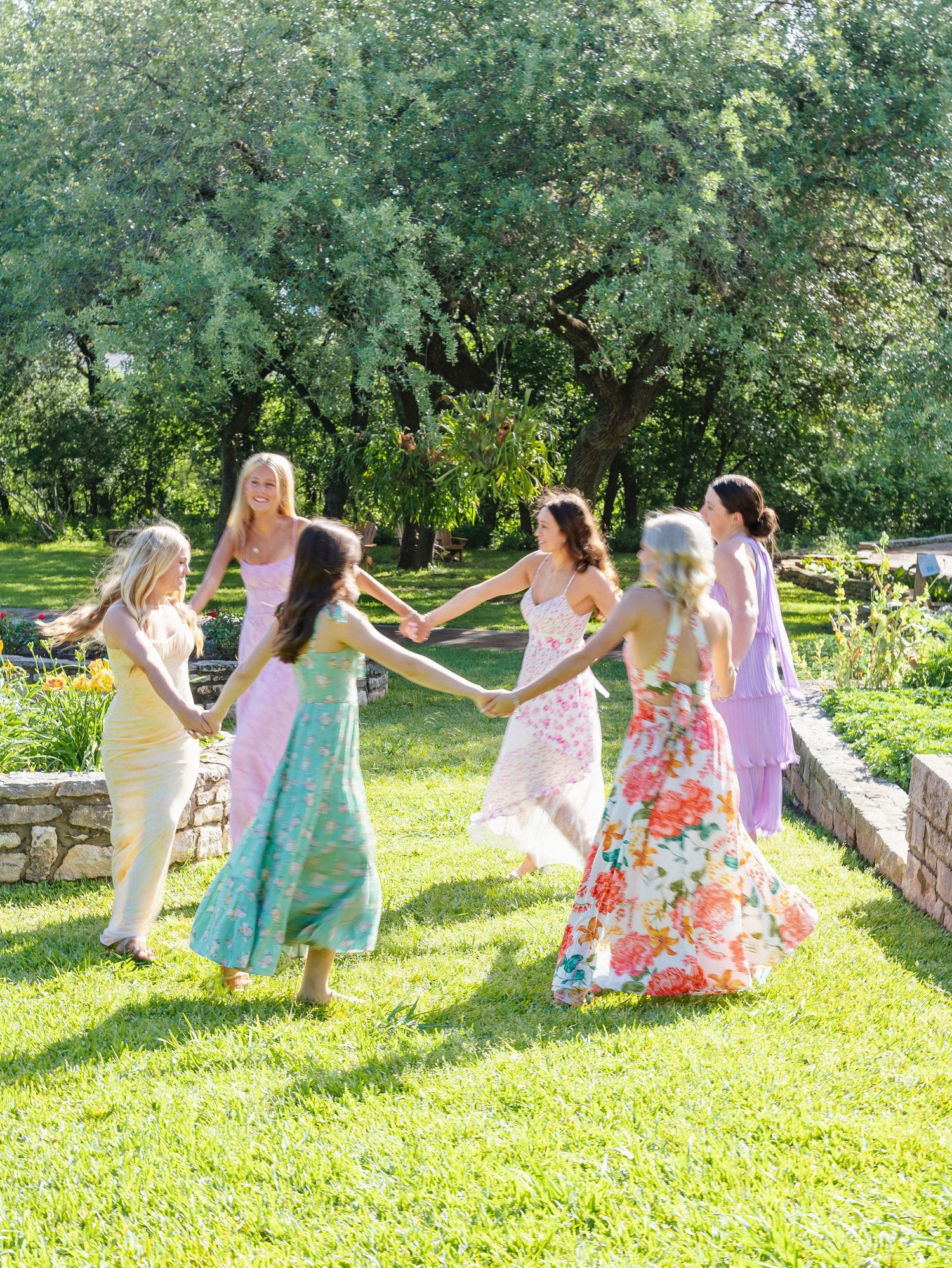 Women in colorful floral dresses holding hands in a circle on green grass surrounded by trees.