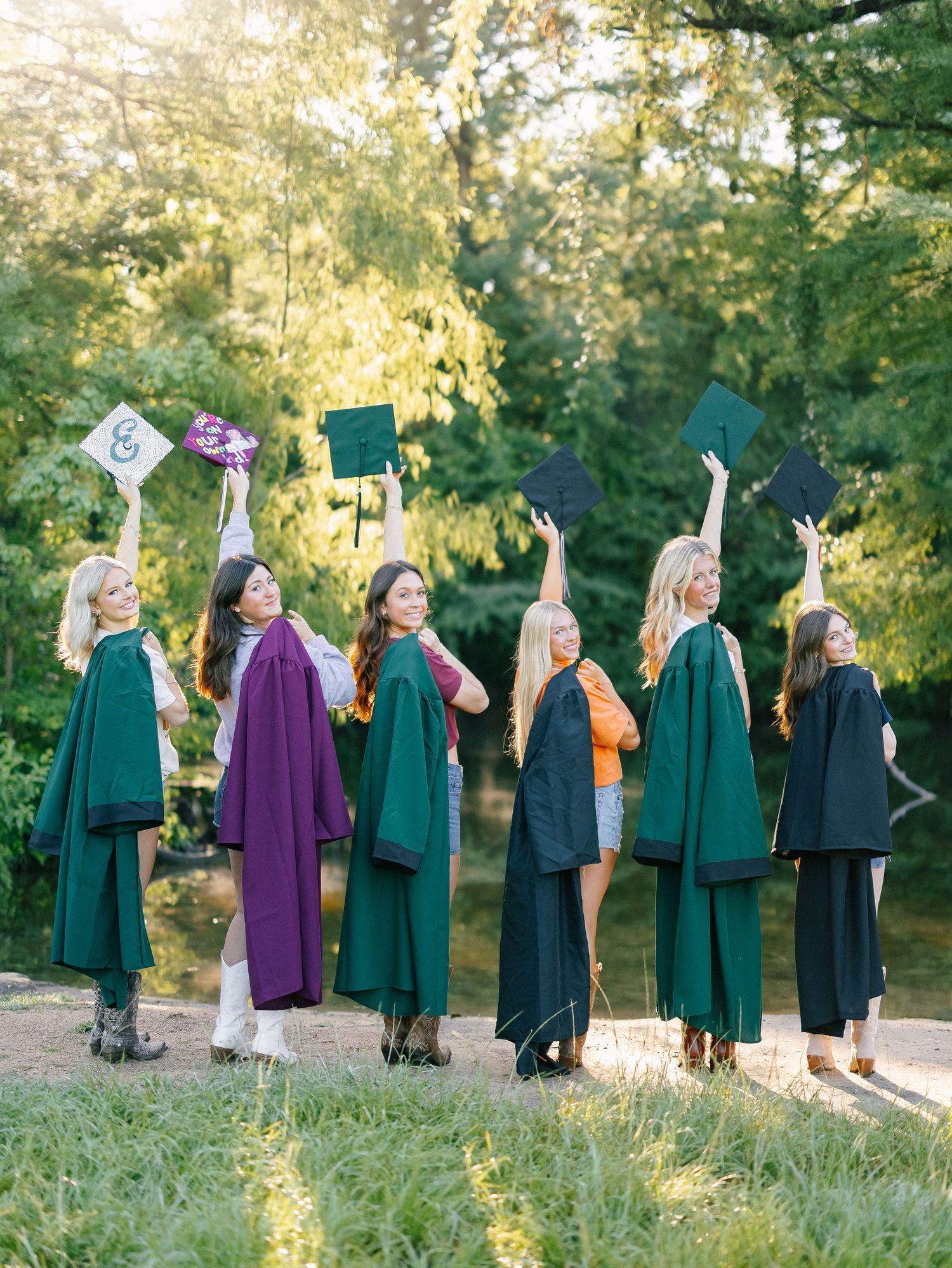 Six graduates in green gowns raise their caps outdoors, celebrating their graduation in a sunny, tree-lined setting.