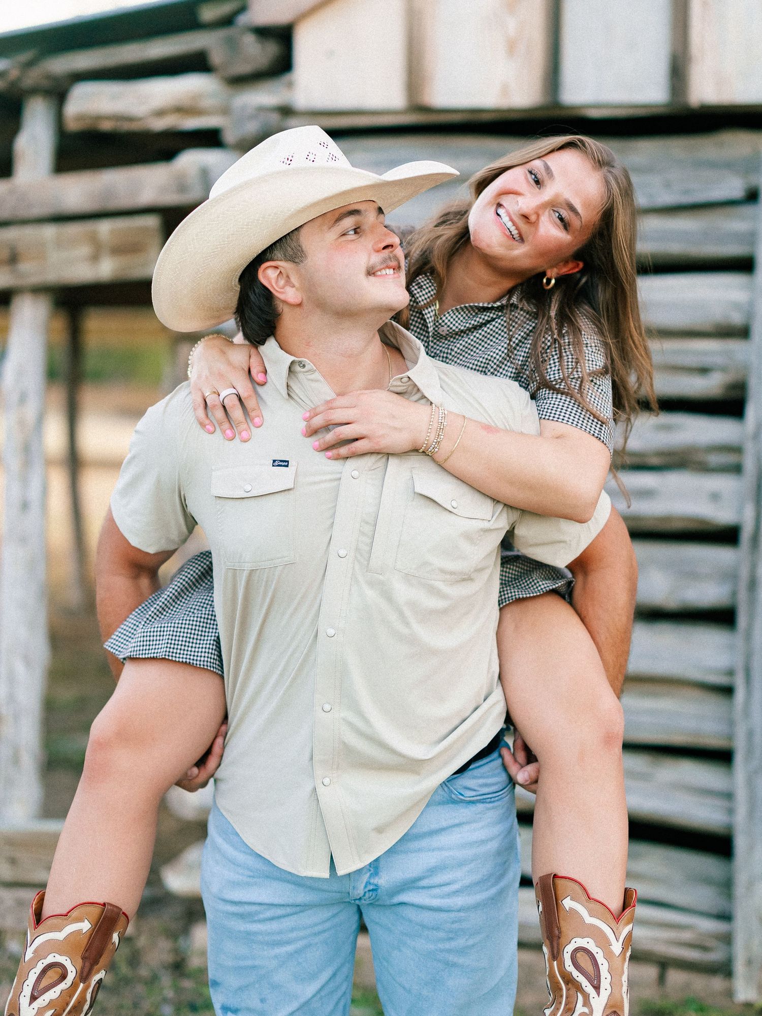 Couple in cowboy attire sharing a piggyback ride outdoors, woman laughing joyfully near a rustic wooden fence.