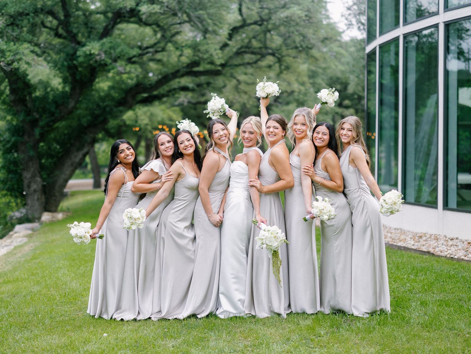 Bride and bridesmaids in silver gowns holding white bouquets, smiling outdoors near lush trees and a glass building.