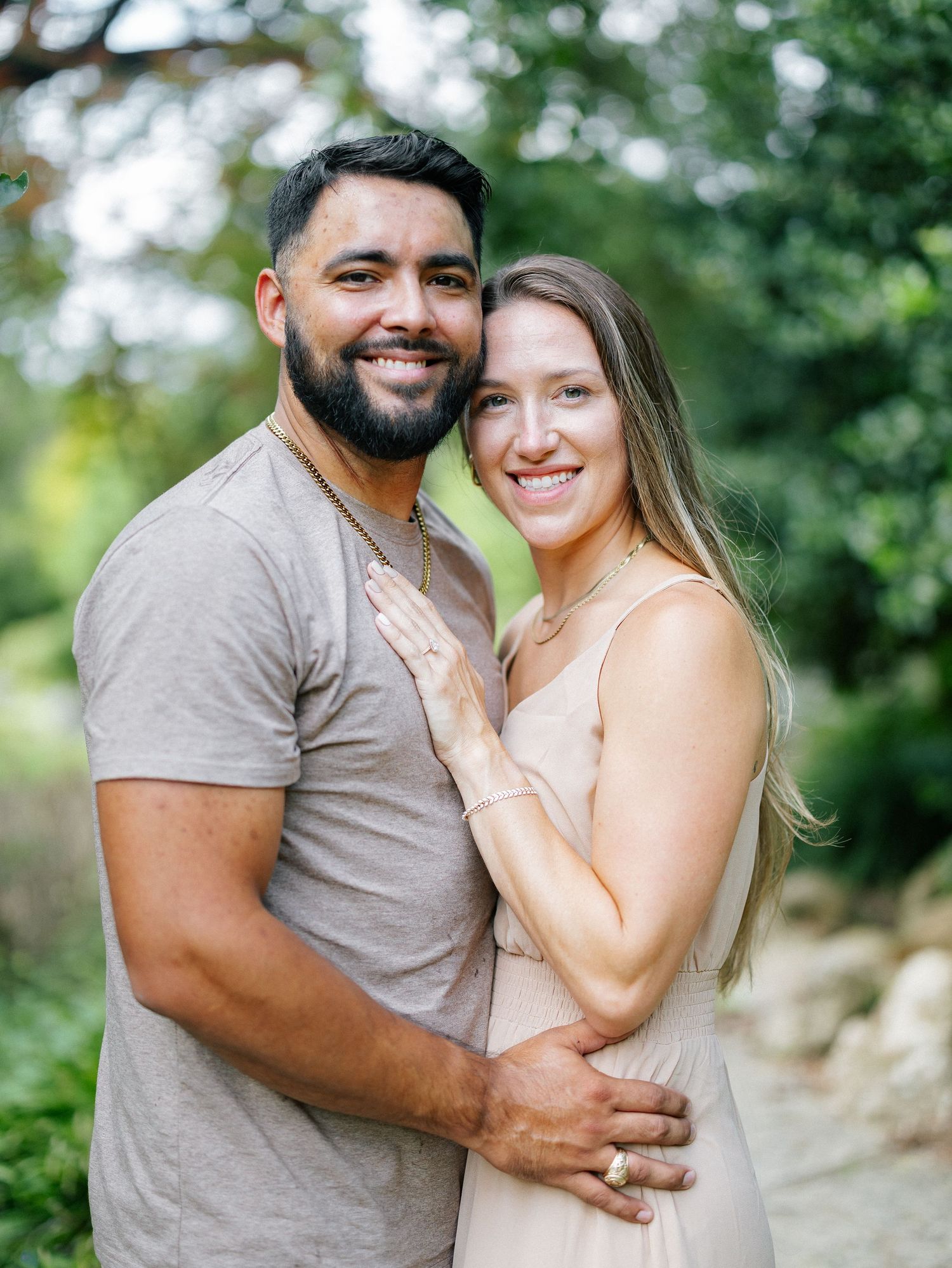 A smiling couple poses closely together outdoors in a natural green garden setting.
