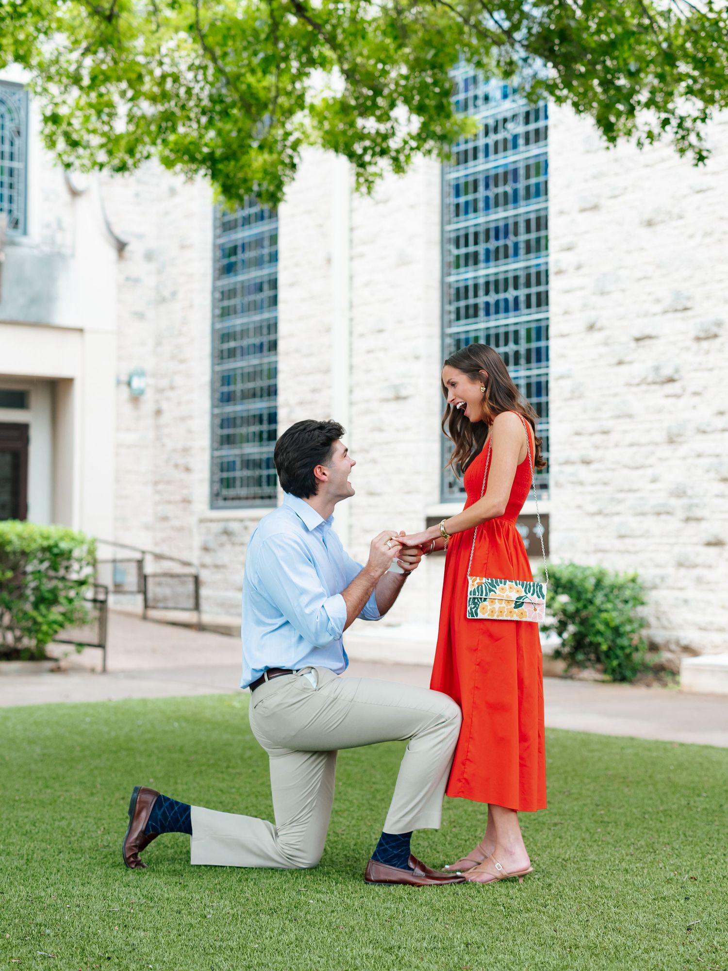 Man proposing on one knee to woman in red dress on green lawn outside stone building.
