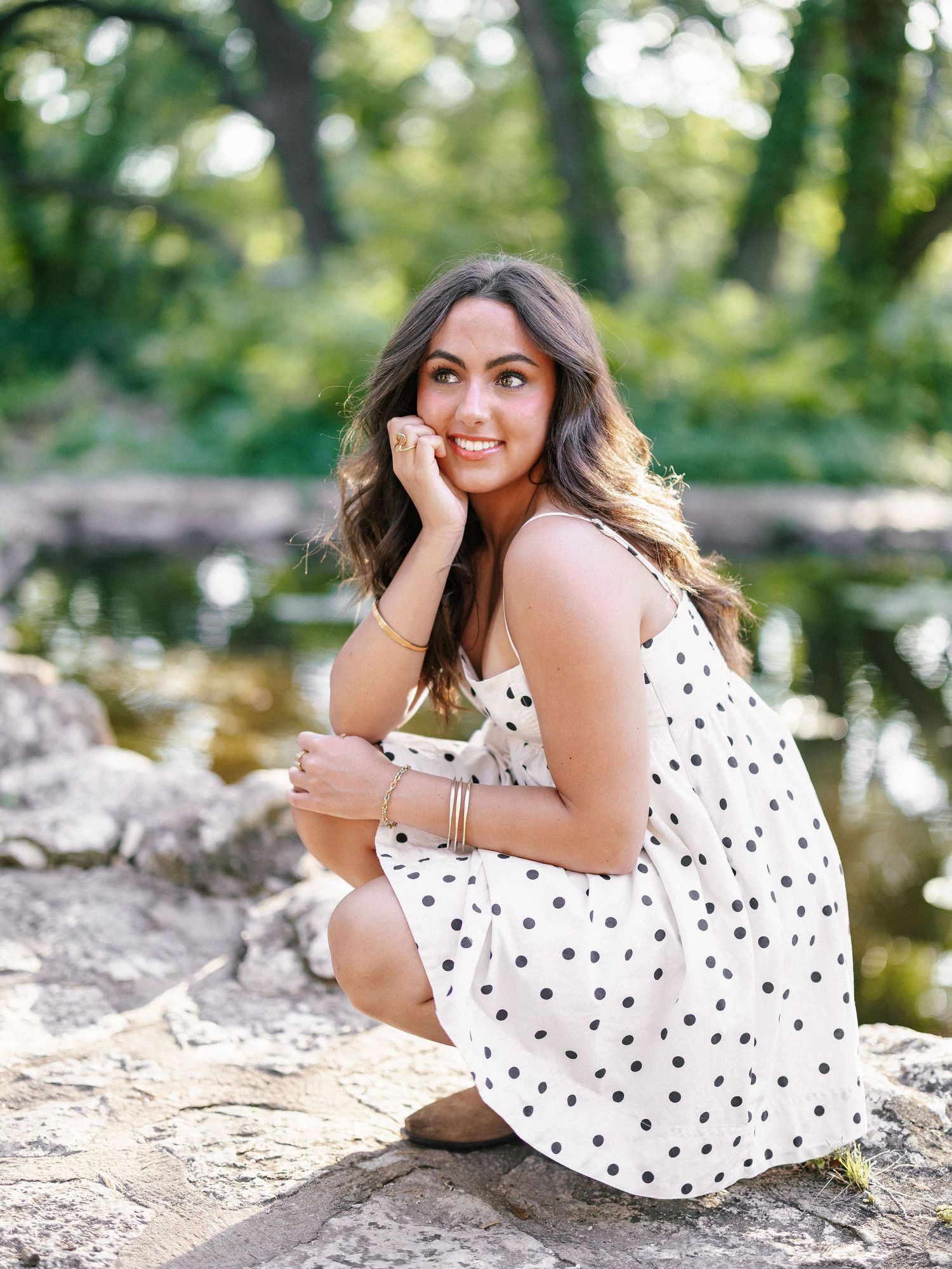 A young woman in a white polka dot dress sits on rocks near a reflective pond in a lush green park.
