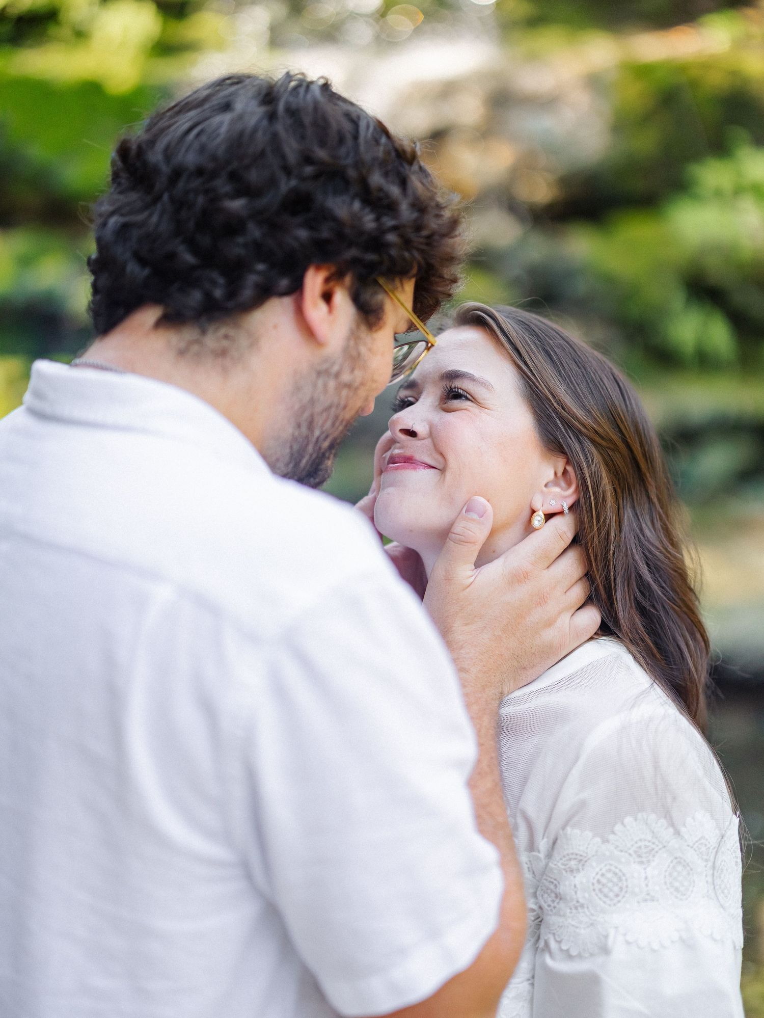 A couple shares a tender romantic moment outdoors, with the man gently holding the woman's face as she smiles up at him.