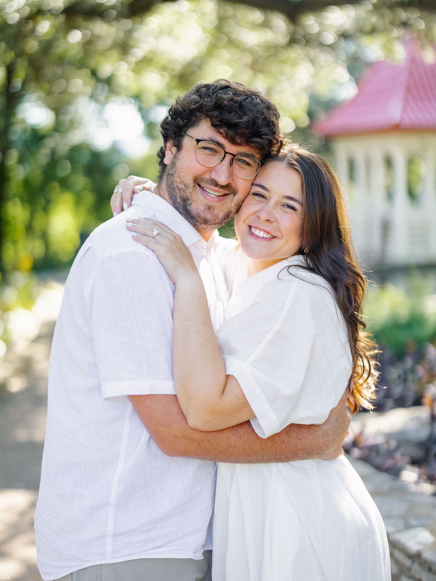 Happy couple in white outfits embracing outdoors, smiling, with lush green trees and a red-roofed building in background.