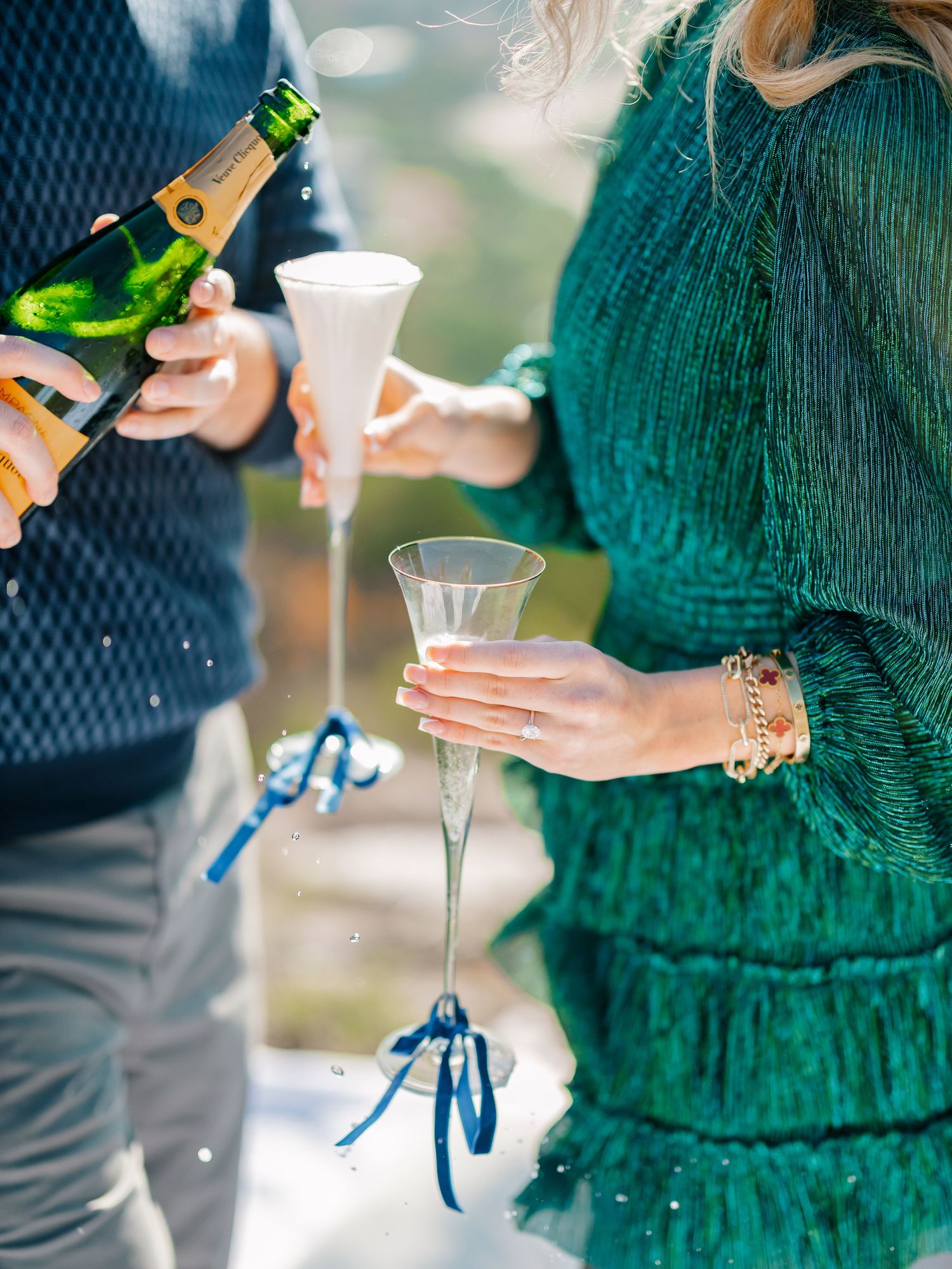 Champagne being poured into crystal flutes tied with blue ribbons at an outdoor celebration.