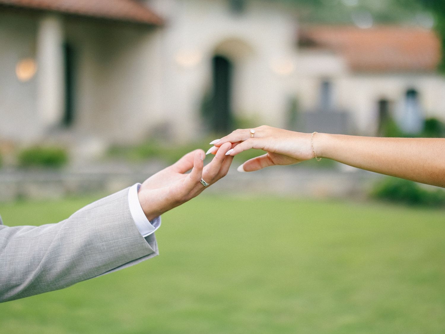 Two people in formal attire gently touching fingertips outdoors near an elegant building, symbolizing a romantic wedding moment.