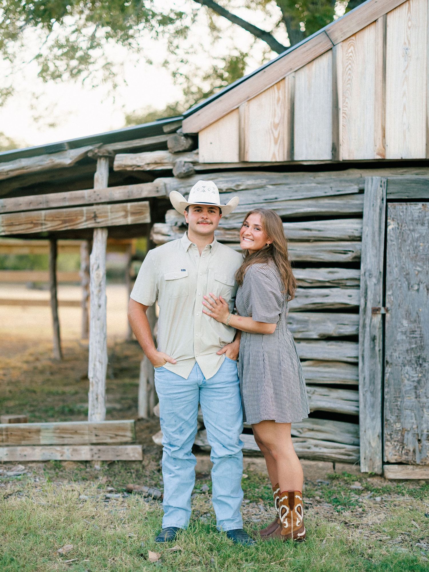A couple in Western attire poses by a rustic log barn, man in cowboy hat and woman in cowboy boots.