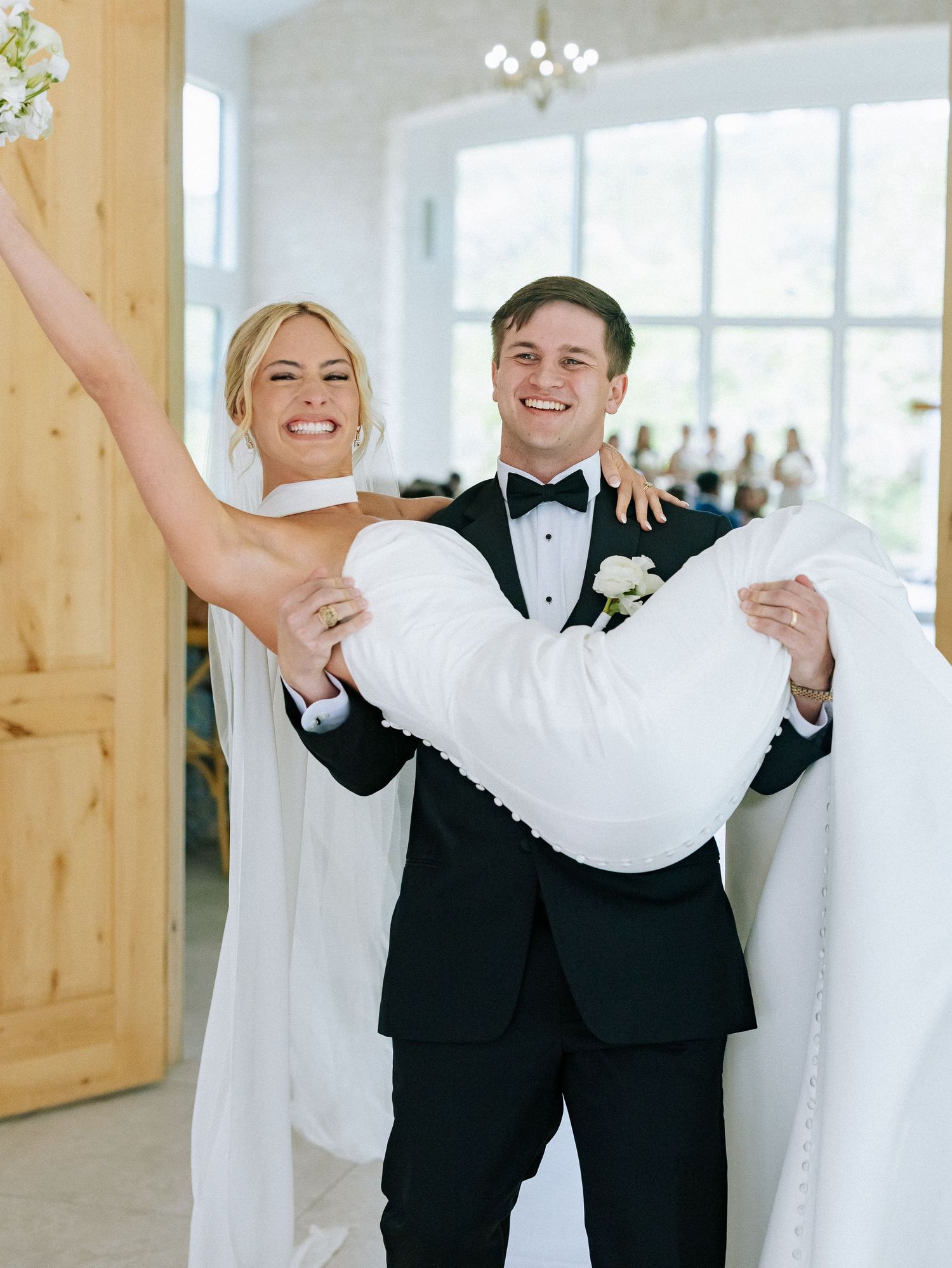Groom in black tuxedo carries smiling blonde bride in white wedding dress as they make their grand entrance.