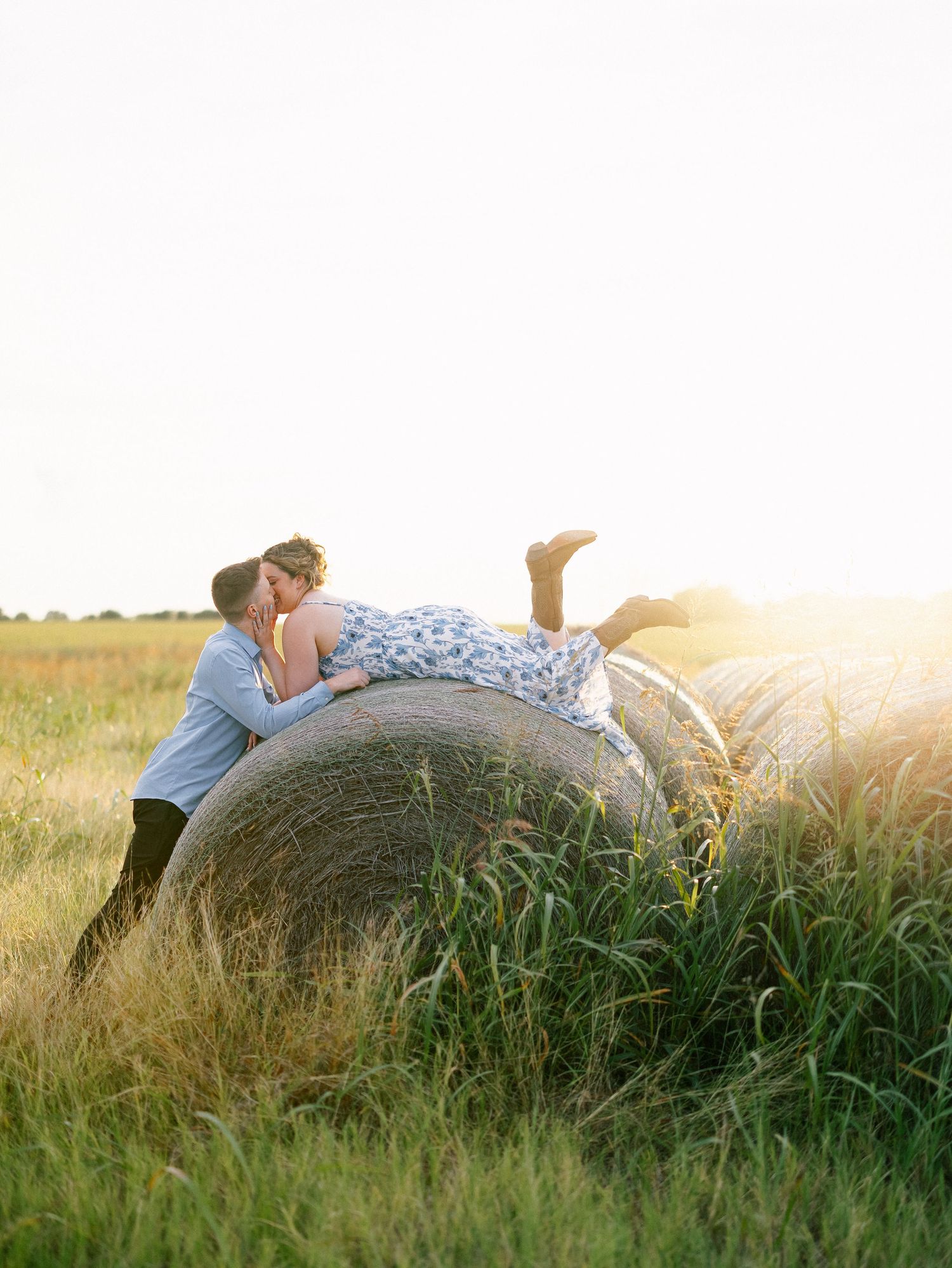 Couple sharing a romantic kiss on a round hay bale in a golden sunlit field during sunset.