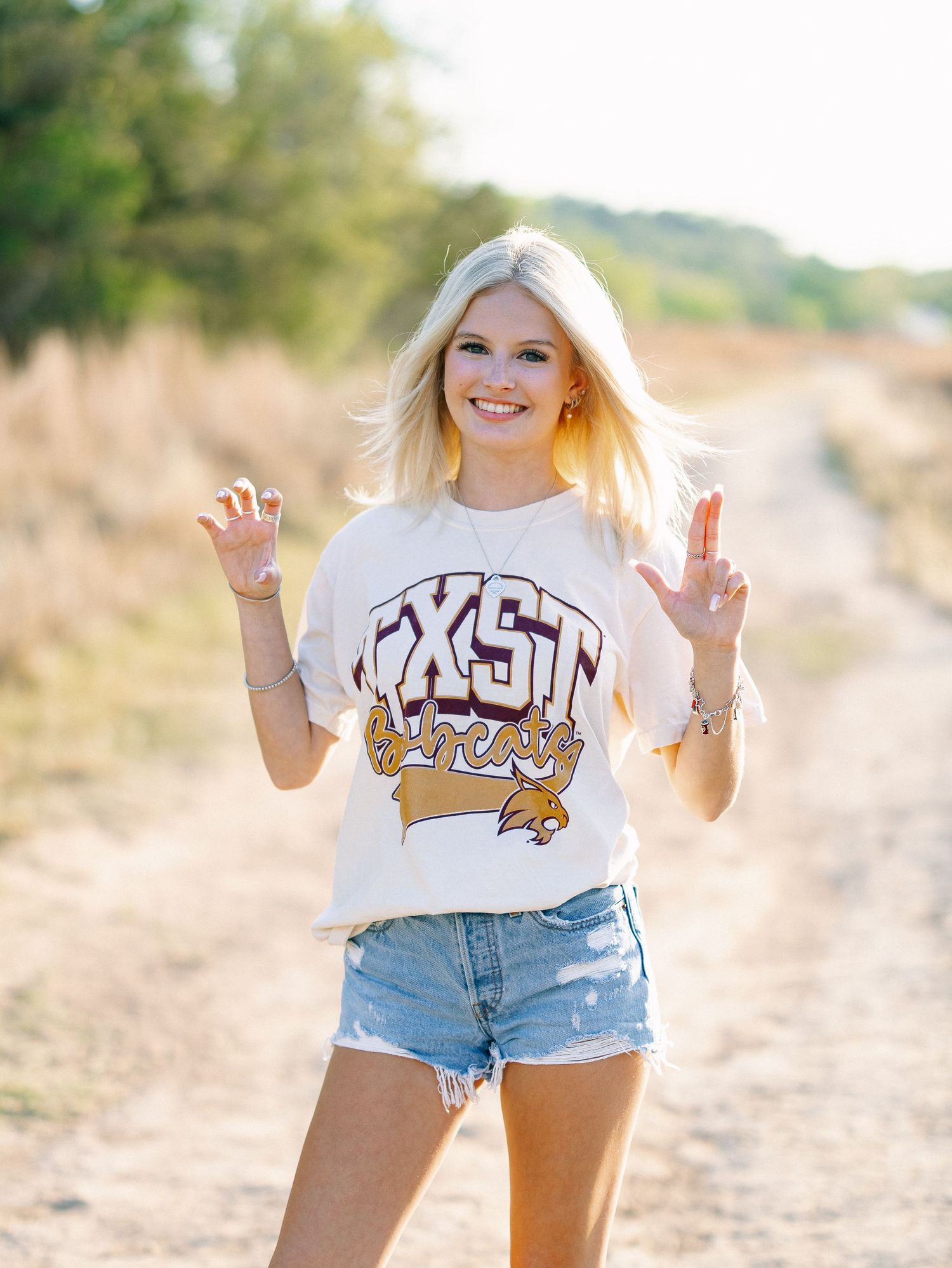 Smiling blonde teen in TXST Athletics tee and denim shorts poses on a dirt path for senior photos.