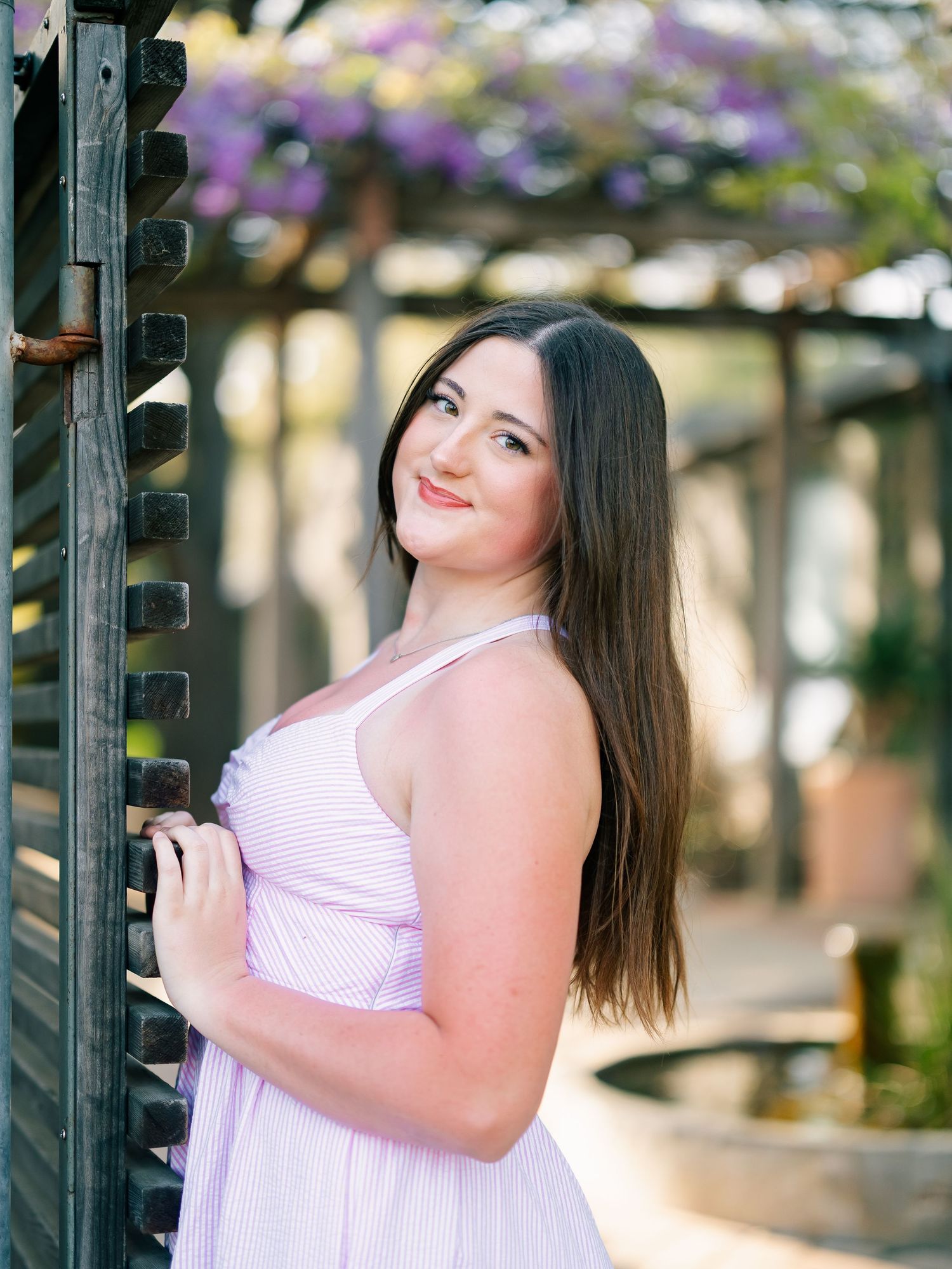 Young woman in lavender striped sundress poses by wooden gate with purple wisteria blooming in background.