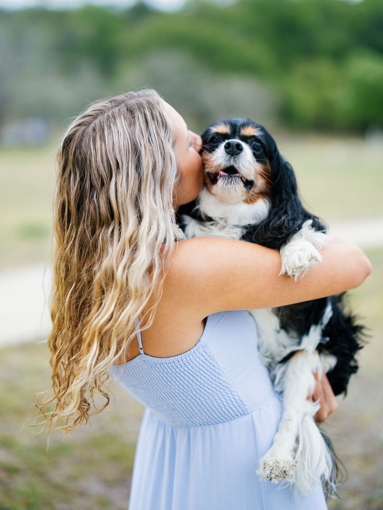 A woman in a light blue dress kisses her Cavalier King Charles Spaniel outdoors.