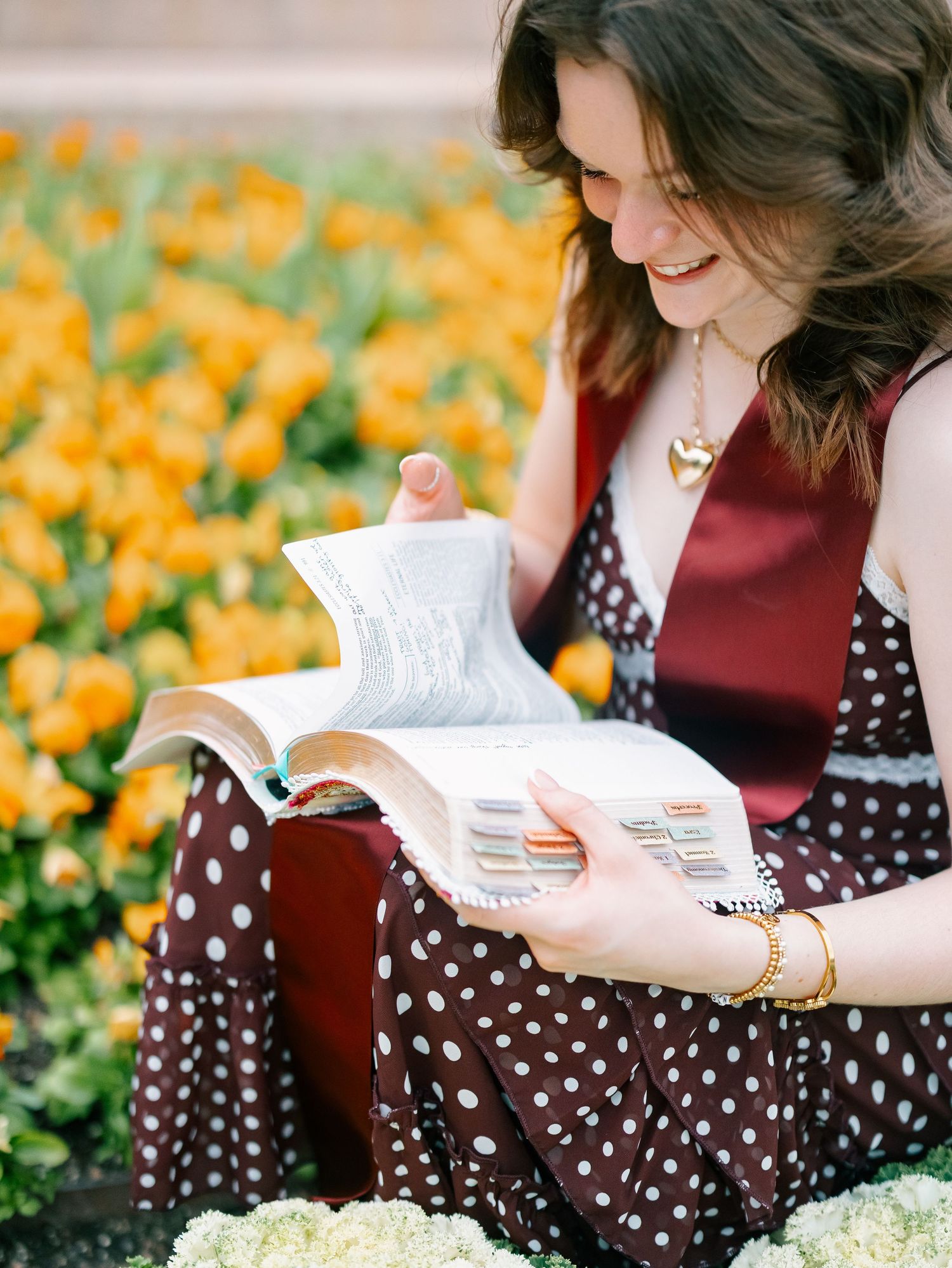 Smiling young woman in polka dot dress flipping through a book beside vibrant yellow tulip flowers.