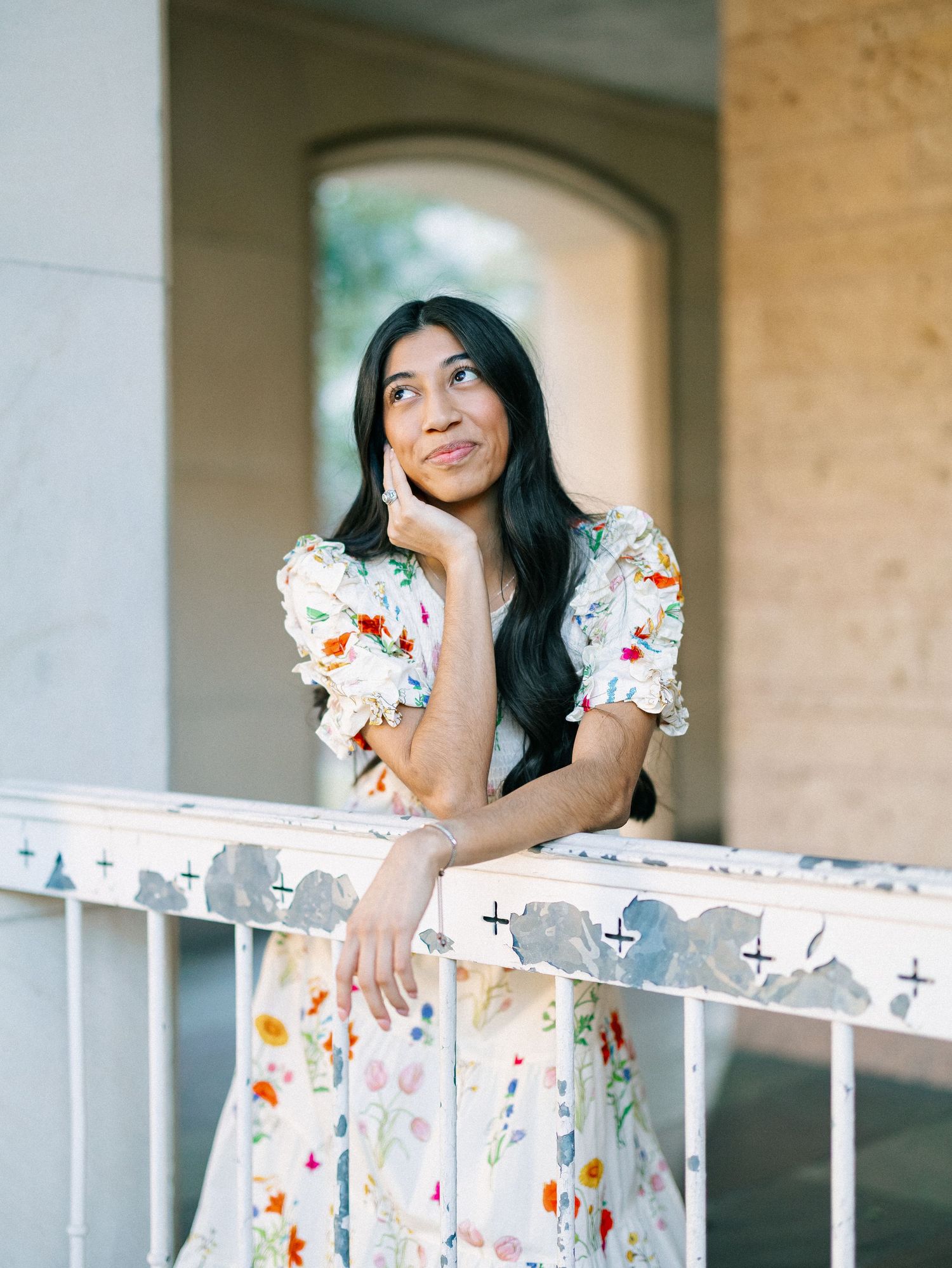 Young woman in floral dress leaning on white railing, gazing upward thoughtfully at an elegant outdoor venue.