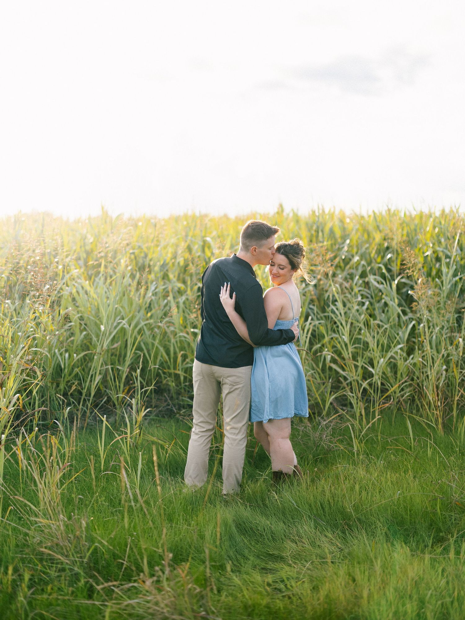 Couple embracing in front of a lush cornfield at golden hour, woman in blue dress looks back softly.