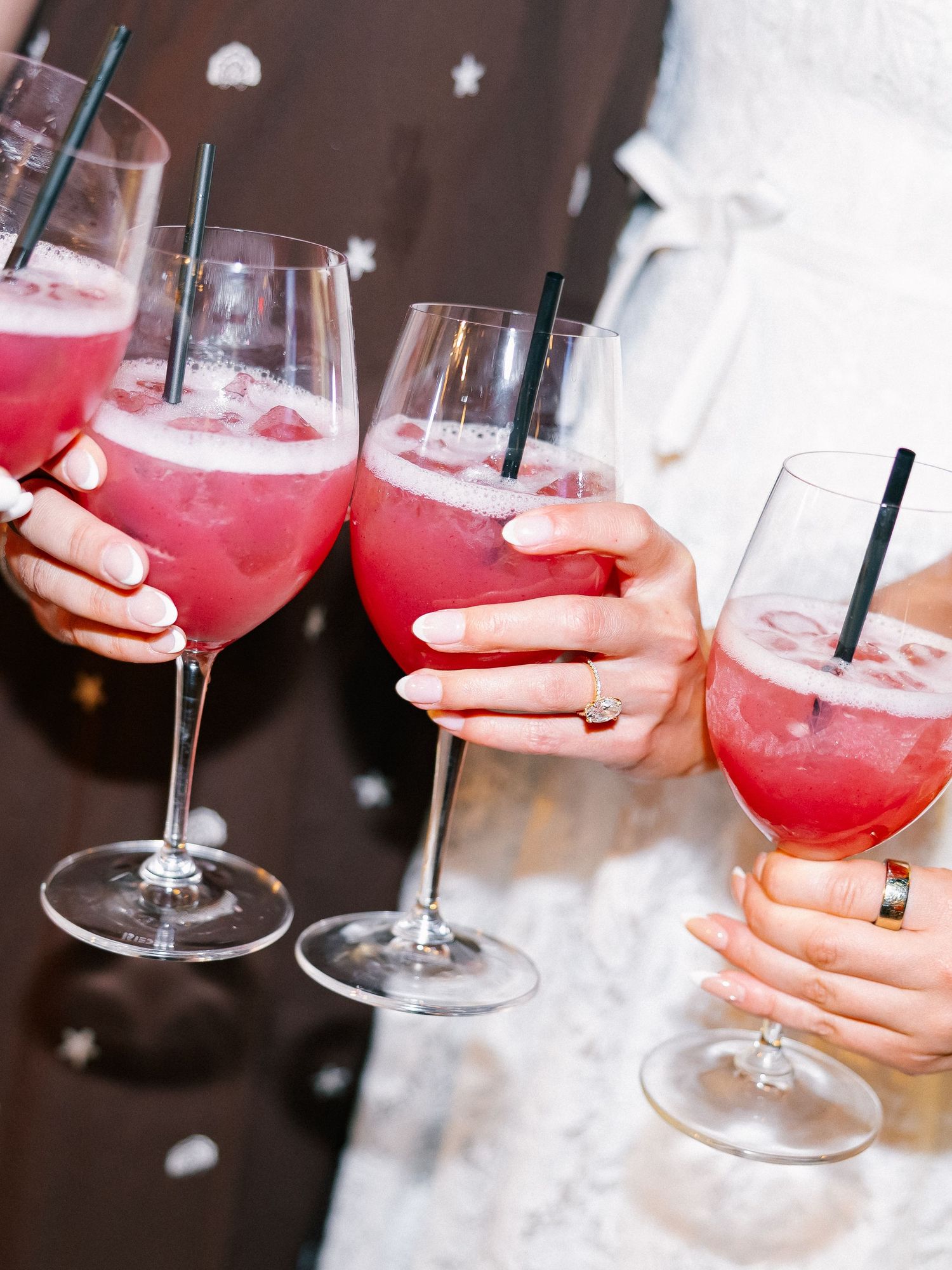 Hands holding pink cocktails in wine glasses with black straws at a wedding celebration.